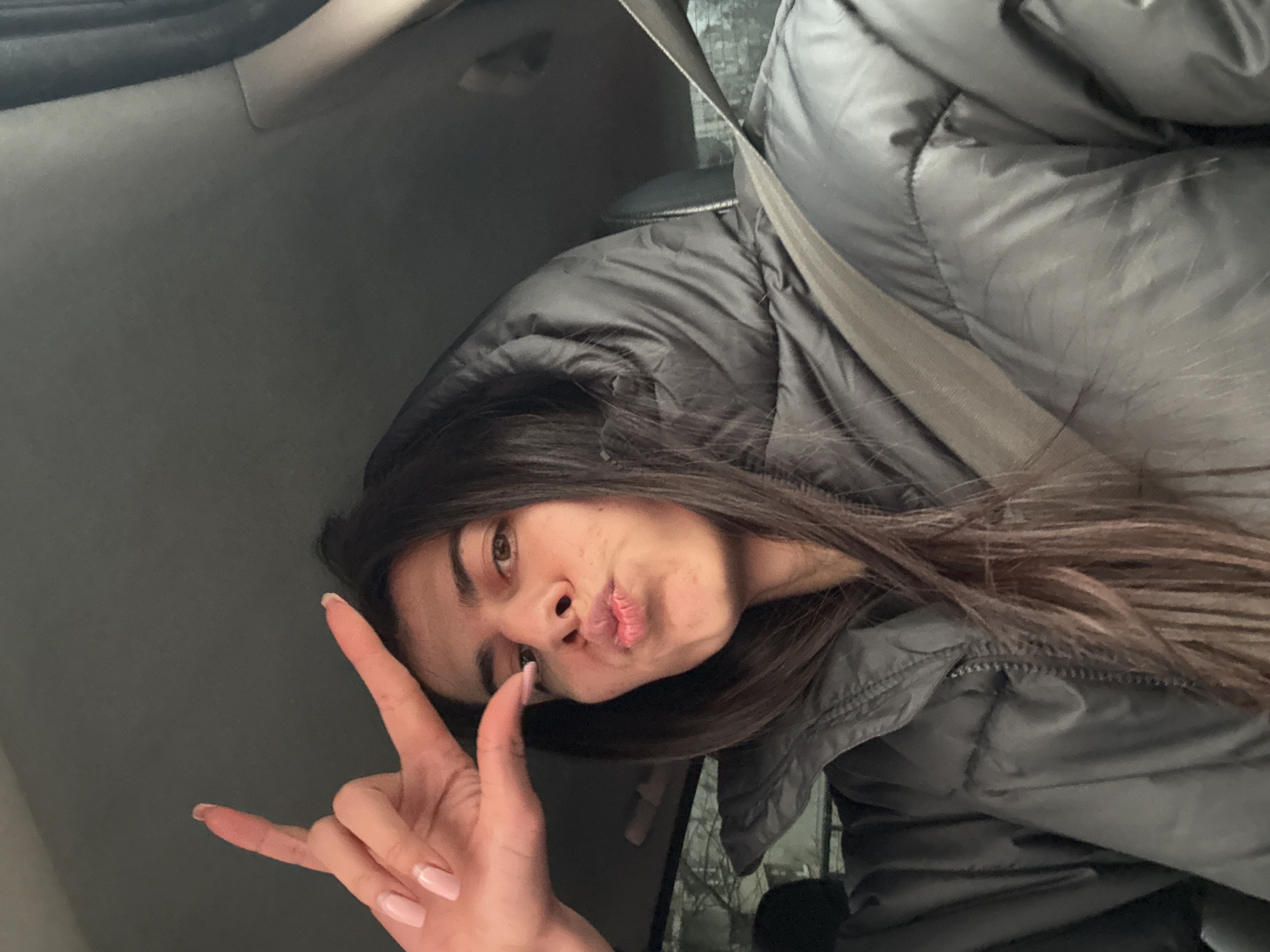 A female individual is depicted within the interior of a vehicle, positioned to face the camera. She has long, dark hair, partially covered by the hood of a dark grey puffer jacket worn over a darker garment. A light-colored seatbelt diagonally crosses her torso from the upper right to the lower left of the frame, indicating she is belted into a seat. Her left hand is raised to the upper left side of her face, forming an 'L' shape with her extended thumb and index finger; her remaining fingers are curled. Her fingernails are manicured with light pink polish. Her facial expression includes pursed lips, forming a "kiss" or "duck face," and her eyes are open, directed towards the viewer. The vehicle's interior features a light grey headliner. Through the window on the left side of the frame, a blurred exterior is visible, suggesting an urban environment with sparse tree branches and building outlines. The precise location within Sofia, Bulgaria, is not discernible from the provided visual information.