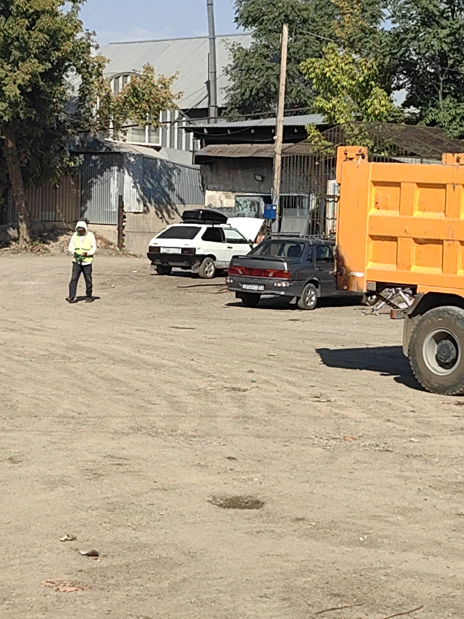 The image depicts an outdoor scene on a sunny day, likely in an industrial or construction area within the city of Shymkent, Kazakhstan. In the foreground, a person wearing a hooded jacket and a yellow safety vest is walking across a dirt lot. The person is holding what appears to be a small electronic device or clipboard.
Behind the person, several vehicles are parked. To the left, a white sedan with a roof rack is visible. To the right of the white sedan, a dark-colored sedan with its doors open is parked. Partially visible in the far right of the frame is the rear of a large orange dump truck.
The background shows a low-lying building with a corrugated metal fence and some trees. The overall environment appears somewhat unkempt, with a dirt or gravel ground surface and scattered debris. The light suggests it is daytime, with clear skies.