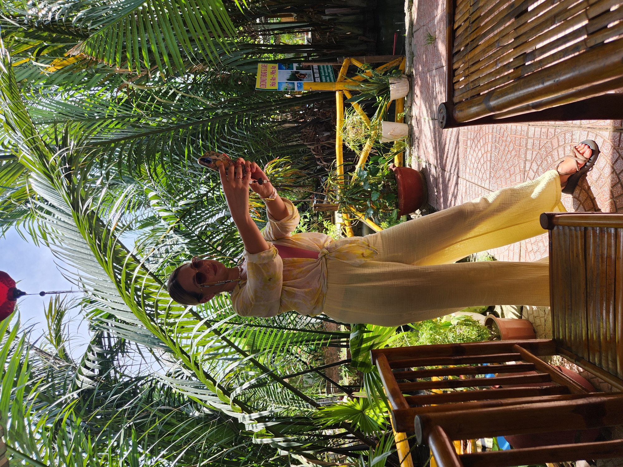 A woman in a yellow tie-front shirt and matching wide-leg pants stands outdoors, holding a cell phone to take a selfie. She wears sunglasses and a necklace. Palm trees and other lush greenery surround her, with a body of water visible in the background. A bamboo table and chairs are in the foreground. A sign that reads "COCONUT BOAT & CAFE Hoi An - Mrs. An" is visible in the background, suggesting the location is in Hoi An, Vietnam, not Da Nang.