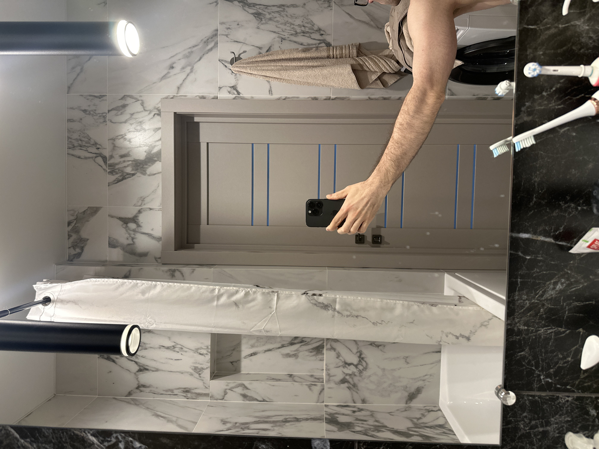 The image appears to be a self-reflection captured in a bathroom mirror.
**Location:** The metadata indicates the location is Kazan, Russia.
**Subjects:**
* A person's arm and torso are visible, holding a smartphone to take the picture in the mirror. A towel hangs from a hook.
* Toothbrushes, possibly electric, are visible on the right side of the mirror.
**Objects:**
* Smartphone
* Mirror
* Bathroom door or cabinet with vertical blue lines
* Towel
* Toothbrushes
* Bathroom tile (both marble and dark, possibly granite)
* Light fixture
* Shower curtain and rod
* Sink
**Actions:**
* A person is taking a photo of themselves in the bathroom mirror.
**Environment:**
* The scene is a bathroom, evidenced by the presence of a shower curtain, sink, and bathroom fixtures. The bathroom features marble tiling on the walls and what appears to be dark granite near the sink area. There is a built-in shelf within the shower area.