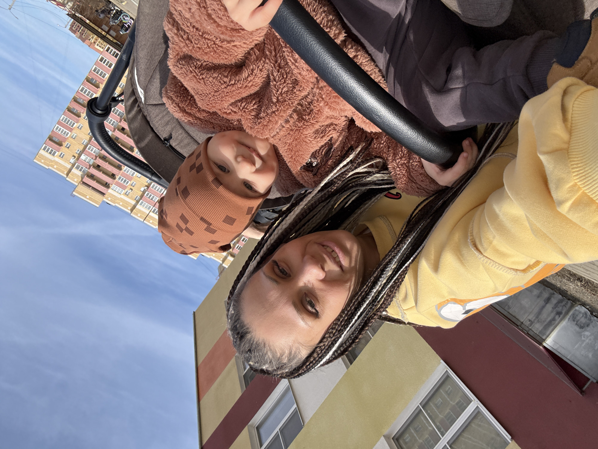 A mother and child are photographed outdoors, possibly near a residential area. The mother, wearing a yellow hoodie, is positioned at the bottom of the frame, smiling at the camera. Her hair is styled in braids with blonde and brown strands. Above her and slightly left, a young child in a brown patterned hat and a furry brown jacket is also looking at the camera and holding onto a black stroller bar. The background shows a low-rise building with a beige and red striped facade, suggesting an urban environment. The sky is visible in the upper left corner of the frame and is a clear blue. In the background, a multi-story residential building with yellow and pink accents can be seen. The specific location is not explicitly stated but is possibly in an urban area, and could be assumed to be in Yaroslavl', Russia.