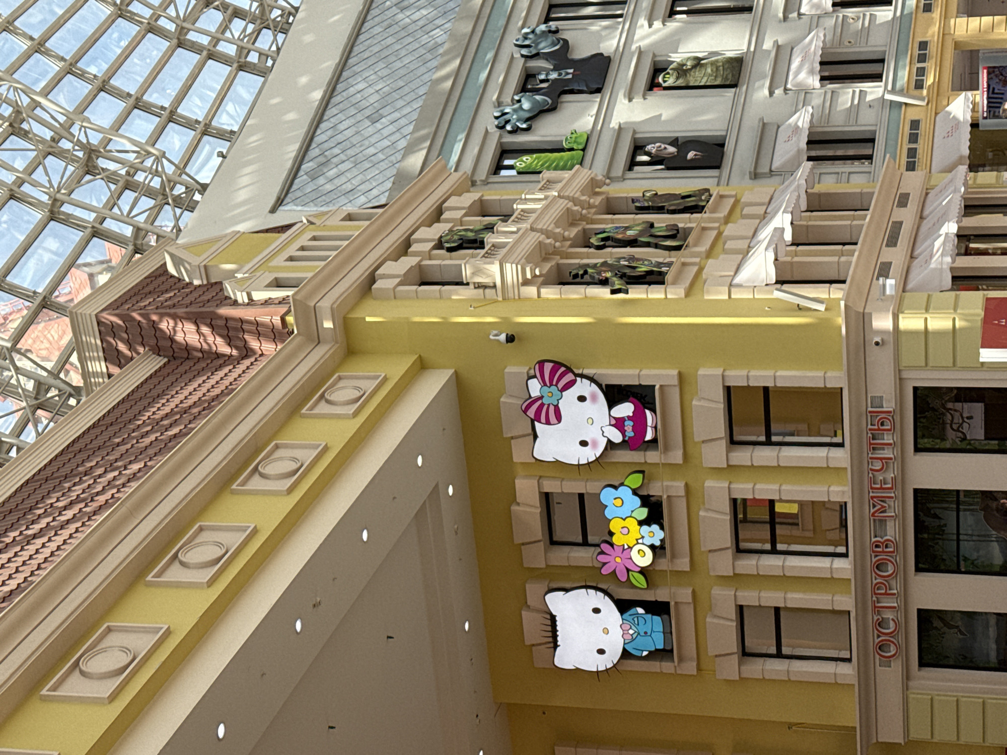 The image shows an interior shot of a building, specifically a section of a large structure likely in Moscow, Russia, as the inscription "ОСТРОВ МЕЧТЫ" (Island of Dreams) is visible, referring to the "Dream Island" theme park in Moscow. The architecture is whimsical, featuring faux building facades with decorative elements.
The building's interior is designed with intricate details, including architectural features that resemble multi-story building sections. There are roof tiles at different levels and decorative windows.
On the walls, there are cartoonish figures like "Hello Kitty" in various poses, adding a playful and fantasy-like atmosphere. Above the cartoon figures, the architectural elements become more complex, including miniature vehicles or figures seemingly peeking out of windows and ledges.
The building has a glass and steel structure that is the dome above.
