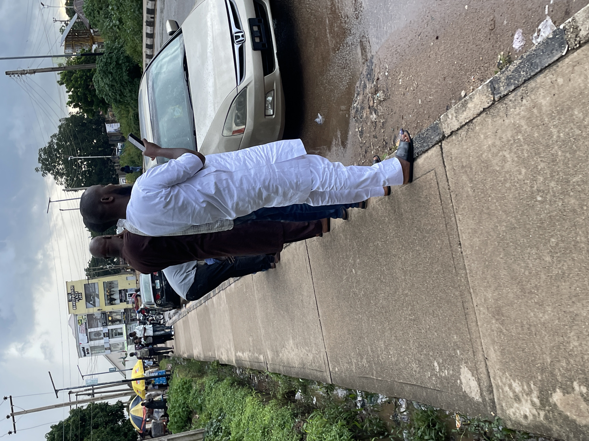 A group of people stand on a concrete sidewalk next to a street in Jos, Nigeria. The street is wet, indicating it has recently rained. A beige sedan is parked on the street. Several people are dressed in traditional Islamic attire, including long robes and head coverings. One man in a white robe is holding a smartphone. In the background, there are buildings, trees, and utility poles. One building has the text "BEAUTIFUL BRIDE PLAZA JOS" on it. There are also yellow and blue umbrellas visible, suggesting a marketplace or street vendors. The scene appears to be a typical urban setting in Jos.