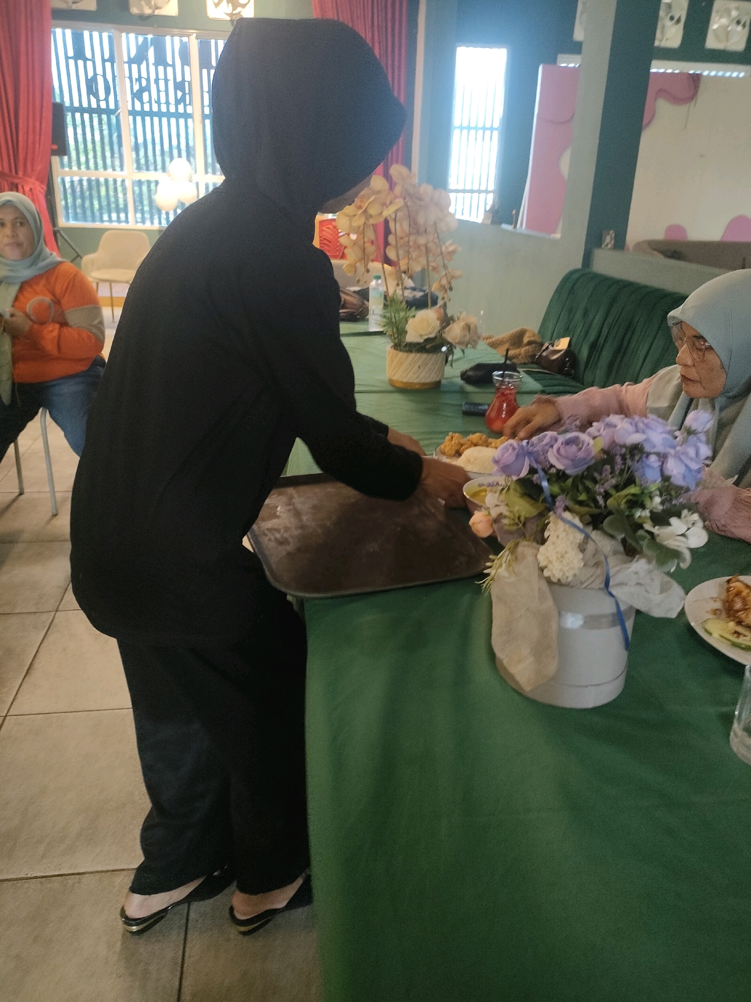 The image captures a moment indoors, likely at a restaurant or event venue in Pekanbaru, Indonesia, suggested by the cultural attire. A person wearing a black hijab and a black outfit is seen from the side, holding a tray and walking. To the left, another person in an orange shirt and grey hijab is seated. In the background, large windows with vertical blinds and red curtains are visible, along with some decorative elements. The foreground features a green tablecloth on a table laden with food, drinks, and a bouquet of purple and white flowers in a vase. The floor appears to be tiled. The scene depicts a casual gathering or mealtime.