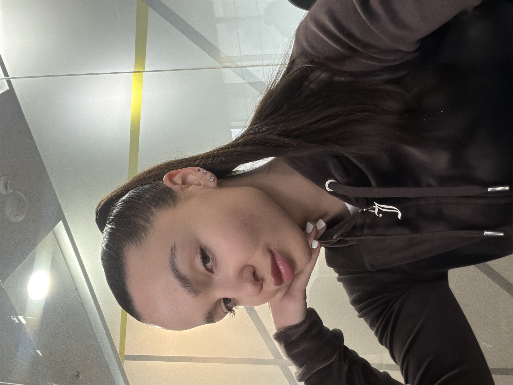 A young woman is captured in a close-up, angled portrait. She is wearing a brown hooded sweatshirt with white drawstrings and a prominent "J" emblem. Her dark hair is styled back from her face, with a ponytail visible. She has multiple piercings in her ear. Her fair skin and dark eyebrows accentuate her features. Her left hand is propping up her chin. The background is a plain, light-colored wall with a distinct yellow stripe. There is no indication in the image that this was taken in Sofia, Bulgaria.