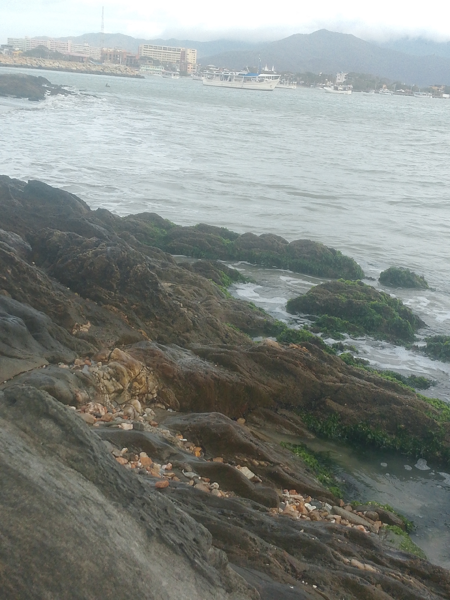 The image shows a rocky shoreline at La Galera in Juan Griego, Venezuela. The foreground is dominated by rocks, some covered in moss and algae, sloping down towards the water. Some loose pebbles and stones are visible on the rocks. The water is gray and choppy, with some white foam from waves. In the background, there are buildings along the coastline and a mountain range obscured by mist or fog. Several boats are visible on the water, anchored or moving slowly. The overall atmosphere is overcast and hazy.