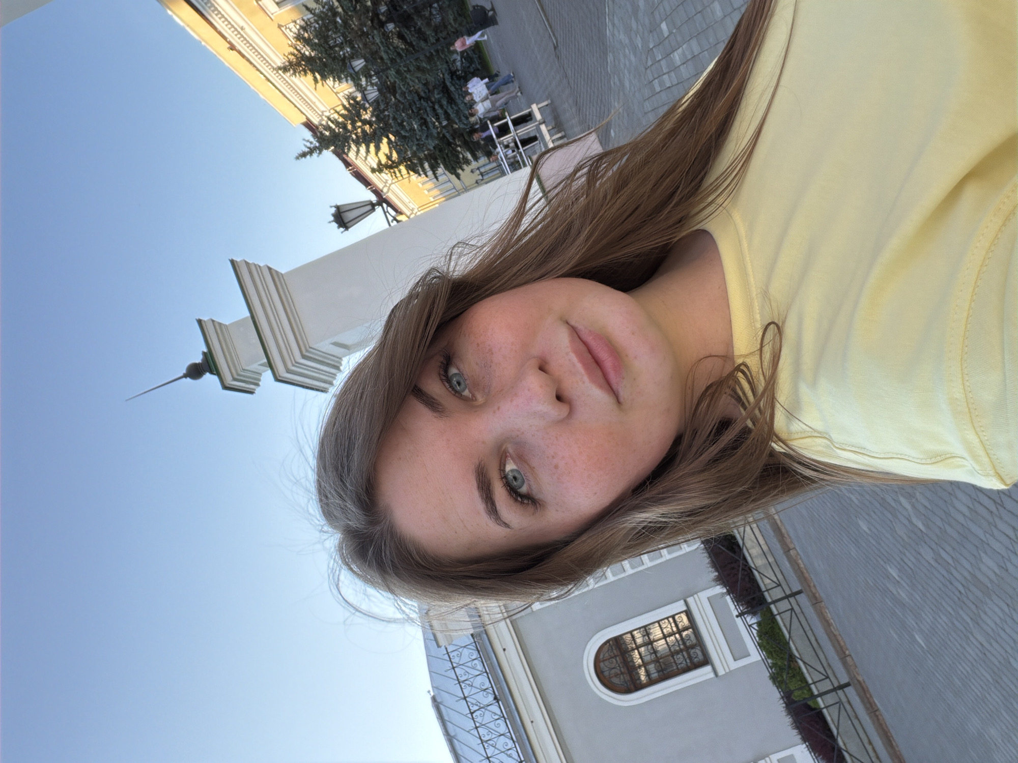 A young female with long brown hair, blue eyes, and visible freckles on her nose and cheeks is depicted in a selfie-style shot, looking directly at the camera with a subtle, neutral expression, wearing a plain light yellow t-shirt. The scene is an outdoor urban environment in Kazan, Russia, under a clear blue sky.
To the left, a light grey building with a dark sloped roof and an arched window with dark grilles is visible, bordered by a black metal fence. In the mid-ground, a white architectural column with green detailing at the top and a dark, pointed spire stands prominently. To the right of this column, an ornate black streetlight is present, and further beyond, a multi-story yellow building with white architectural trim is partially obscured by several dark green coniferous trees. The ground is paved with light grey cobblestones or pavers. In the background on the right, several individuals are walking away from the camera's perspective on the paved street.