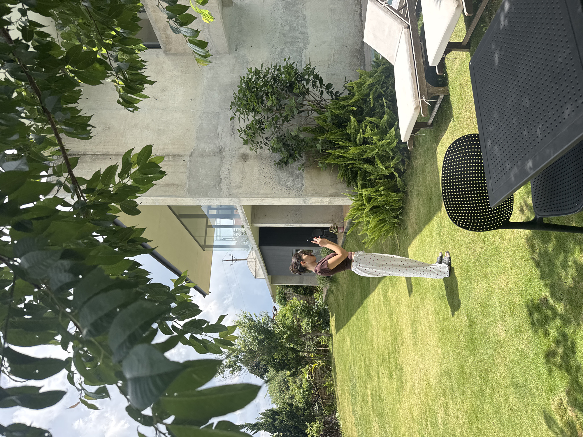 In Đa Lộc, Vietnam, a young woman stands in a lush, green lawn, holding a smartphone to take a photo. She is wearing a brown t-shirt and white pants with small red hearts. Behind her, a modern concrete building with large windows and a sloped roof is partially visible, surrounded by greenery including trees and ferns. Two lounge chairs with beige cushions are placed on the grass in the foreground, along with a black outdoor table and chairs. The sky is cloudy.