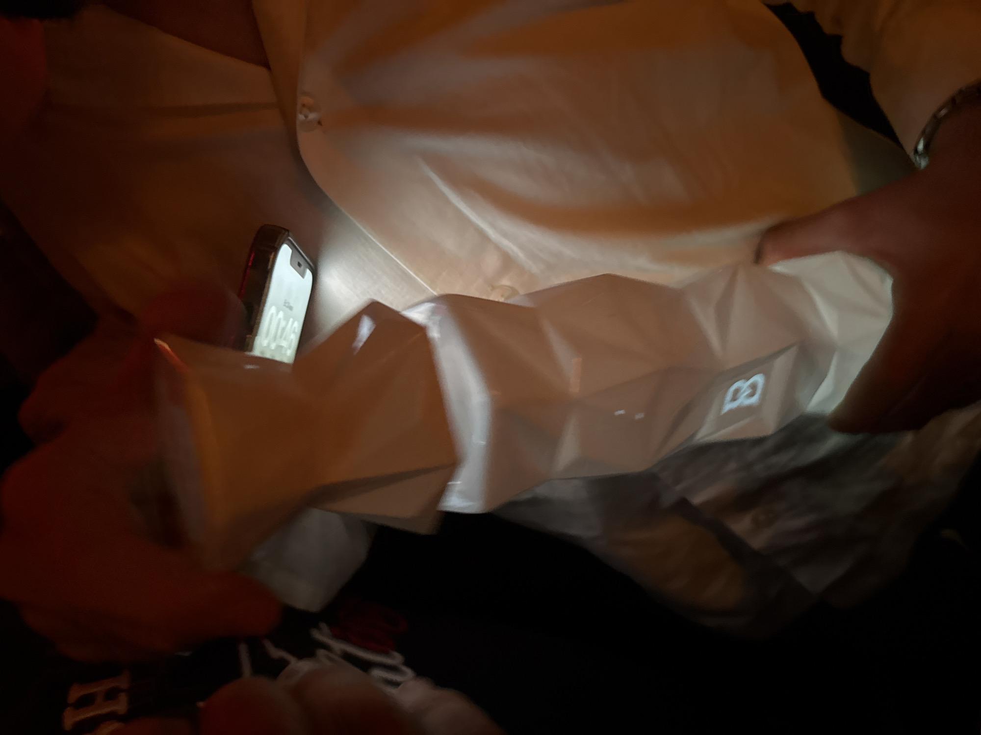 A person illuminates the inside of a white, angular shopping bag with a smartphone. The bag features a visible "B" logo. The person is wearing a light-colored shirt. The location is Yerevan, Armenia.