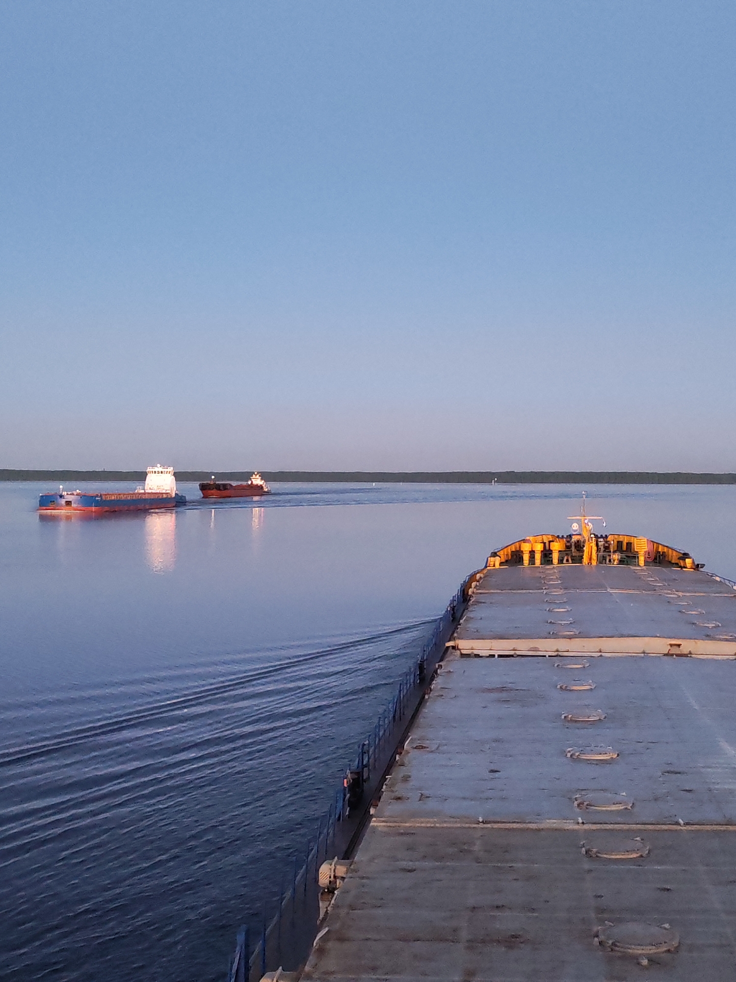 The image captures a maritime scene on a wide, calm body of water under a clear, pale blue sky, described as being near Malinukha, Russia. In the immediate foreground, the grey, flat deck of a large vessel, possibly a barge or cargo ship, is observed from a stern perspective. The deck features multiple rows of circular metal hatches and is bordered by blue railings on the port side. Towards the forward end of this vessel, a structure with yellow and orange components is visible. A discernible wake emanates from the vessel's port side.
In the midground, two additional vessels are underway, moving from left to right across the frame. The vessel positioned to the left displays a blue hull with red trim and a prominent white superstructure, indicative of a river push-boat or a self-propelled barge. To its right and slightly astern, a second, darker-hulled cargo ship or tanker, featuring a white superstructure at its stern, is also in motion. Both midground vessels are generating distinct wakes on the water.
A long, linear, dark green landmass, appearing as a distant treeline or low-lying shore, forms the horizon. The lighting suggests either dawn or dusk, with soft illumination reflecting off the calm water surface. No individuals are visible in the scene.