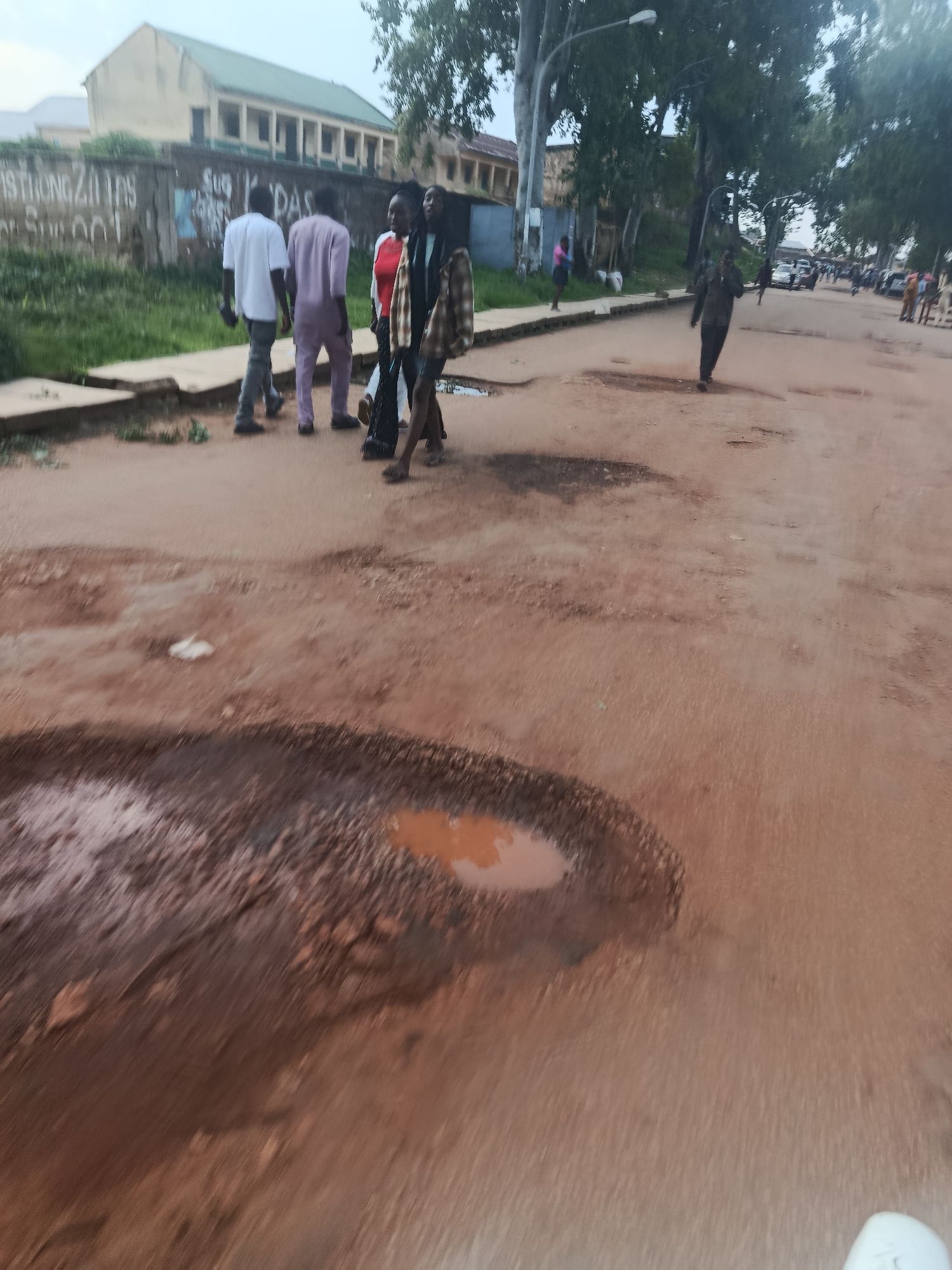 A group of young people are walking on a dirt road in Rafin Par, Nigeria. The road is uneven and has several large potholes filled with muddy water. The people appear to be casually dressed, with some wearing t-shirts and pants, and others in more traditional clothing. The environment is a mix of natural and man-made elements, with trees and vegetation lining the side of the road, as well as buildings in the background. The sky is overcast, suggesting it may have recently rained or is about to. The image is taken from a low angle, giving the impression of movement.