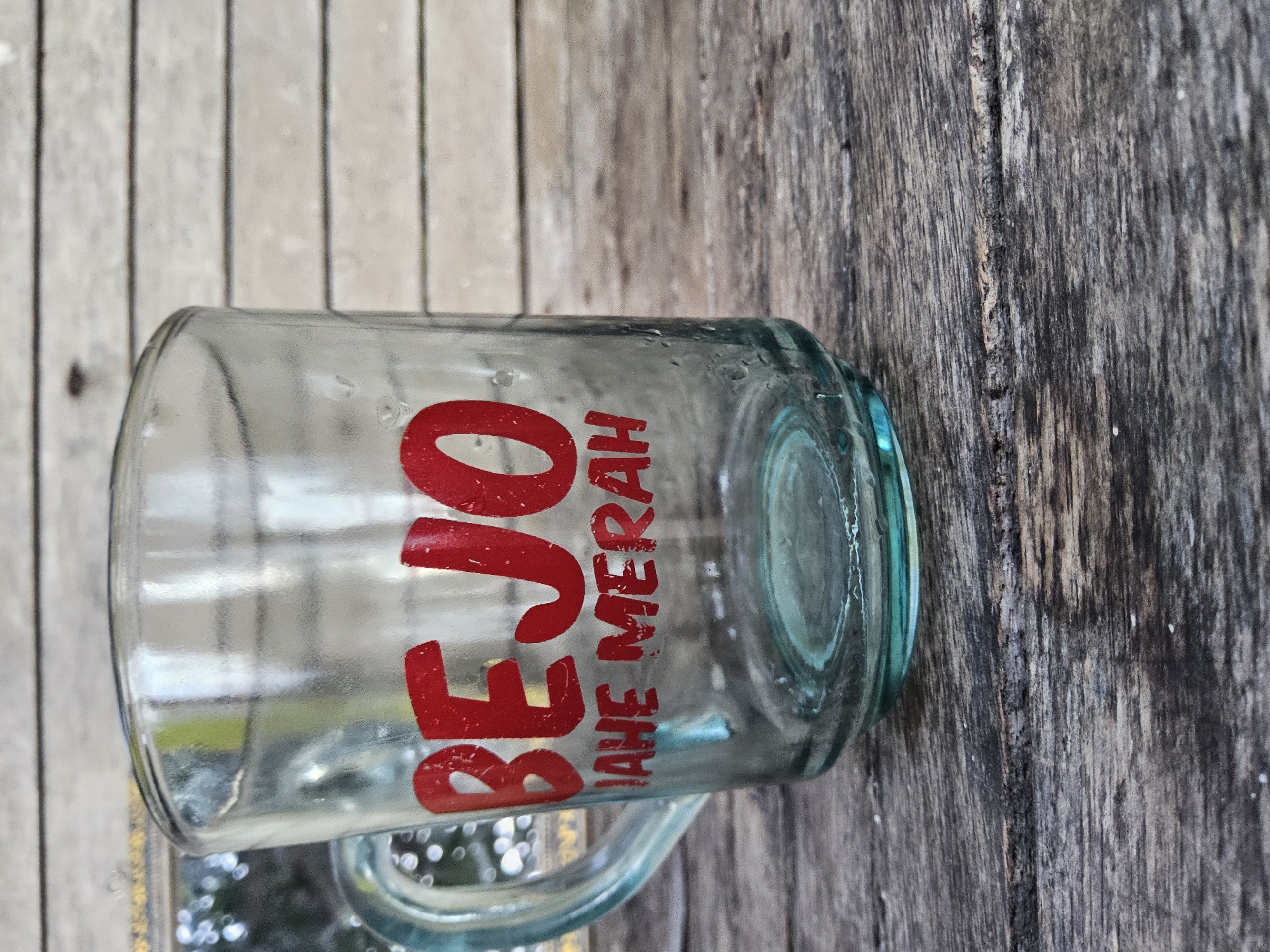 A clear glass mug with red text is placed on a weathered wooden surface. The text on the mug reads "BEJO JAHE MERAH". The mug has a handle and condensation droplets are visible on its exterior. The background consists of horizontal wooden planks, suggesting an outdoor or rustic setting. No people or actions are depicted in the image. The location in the city of Payagaboh, Indonesia is not identifiable from the visual information provided in the image.