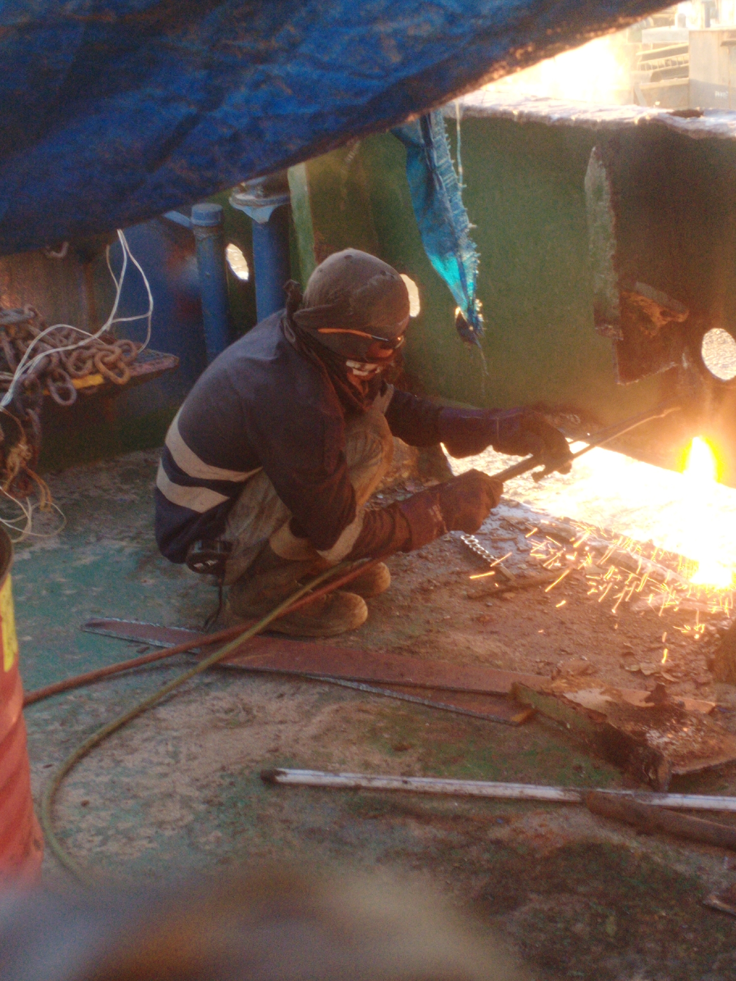 An individual, clad in protective gear including a helmet and gloves, is kneeling on a weathered deck and engaged in a cutting operation with a torch. Sparks are actively flying from the torch's point of contact with a metallic surface, illuminating the immediate area with a bright orange glow. The surrounding environment appears industrial, with visible chains and other machinery components. The lighting suggests an outdoor setting, potentially during daylight hours, though the presence of artificial light from the torch is a dominant feature. The ground is covered in debris and dirt. The image captures a moment of intense activity within a confined or sheltered workspace, possibly on a vessel or in a shipyard. The location in Siddo, Indonesia, suggests a maritime or industrial context.