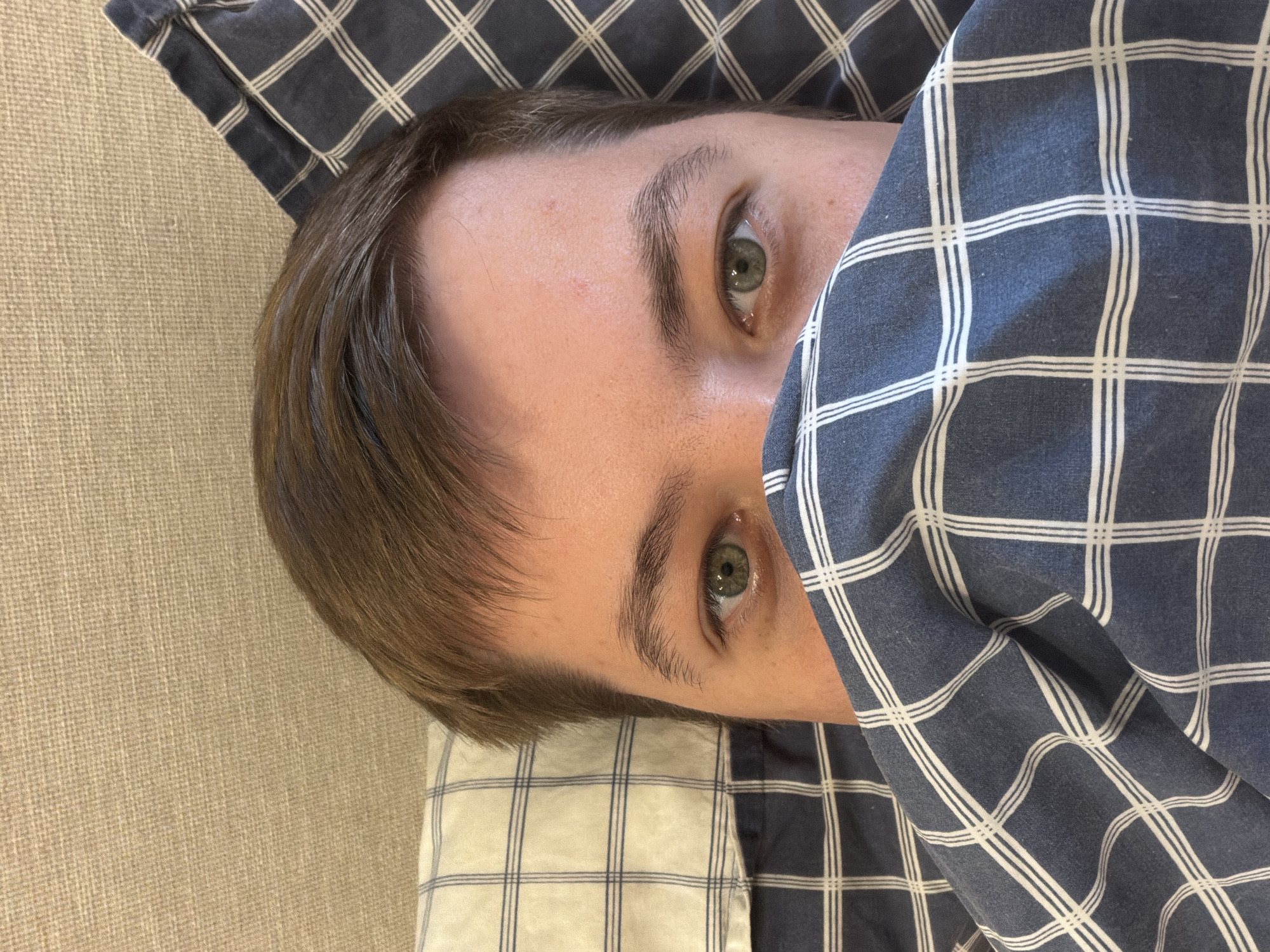 The image, taken in Budva, Montenegro, features a close-up, vertically oriented shot of a person's upper body, mostly their head, partially covered by bedding. The subject is seen from a slightly overhead perspective, lying down.
The person has fair skin, light brown hair, and striking green eyes that are open and looking directly towards the viewer. Their forehead and the right side of their face are visible, while the left side is largely obscured by a blanket. There is a small, faint reddish mark on their forehead. Their hair is short and appears to be swept to one side.
The person's head rests on a pillow with a light blue and white checkered or plaid pattern. They are covered by a darker blue and white plaid blanket, which features thick white lines forming the grid. This blanket is pulled up to just below their eyes, creating a cozy and partially hidden appearance. To the left and top of the frame, a textured, light brown or beige surface, possibly a wall or headboard, forms the background. The overall scene suggests an indoor setting, likely a bedroom, with the person resting or waking up in bed. There is no visible text in the image.