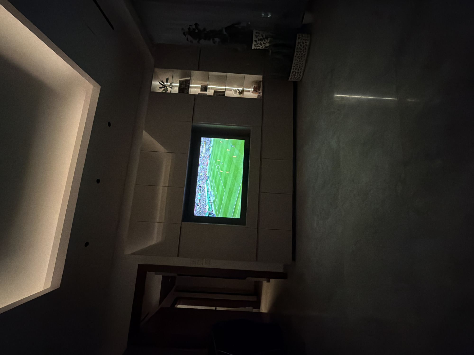 In an indoor living room, a television screen displays a soccer game. The room is dimly lit with recessed ceiling lights providing illumination. The television is mounted on a wall unit with cabinets below. To the right of the television, a tall, narrow display cabinet with shelves and internal lighting showcases decorative items, including plants. A decorative plant in a patterned pot is visible on the floor to the right. The floor appears to be polished stone or concrete, reflecting some of the light. A doorway is visible to the left, leading to another room. The overall scene suggests a modern, contemporary living space.