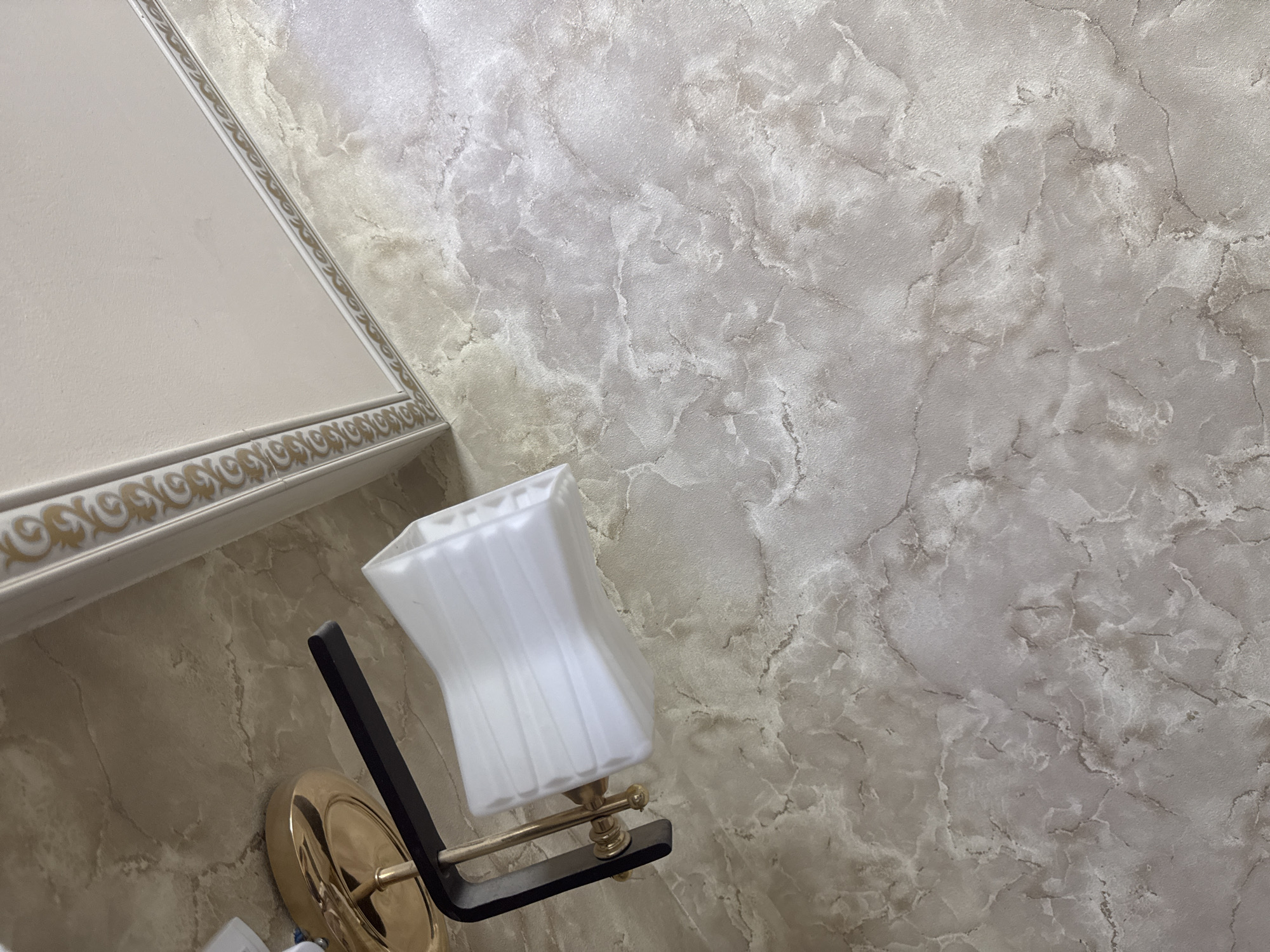 The image shows a wall with a marble-like texture and a portion of a light fixture. The wall is decorated with a marbled pattern in shades of beige and gray, with darker veins running through it. A decorative border with a gold ornamental pattern is visible along the top left edge of the image, suggesting it's part of a cornice or molding. A wall-mounted light fixture with a white, pleated lampshade is positioned on the wall, extending from the left side of the frame. The base of the light fixture appears to be made of brass or gold-toned metal and is attached to a black arm. The overall scene depicts an interior detail, likely within a building. There are no people or obvious signs of human activity in the image. The location in Shofirkon, Uzbekistan, is not visually indicated by any landmarks or contextual clues within the image itself.
