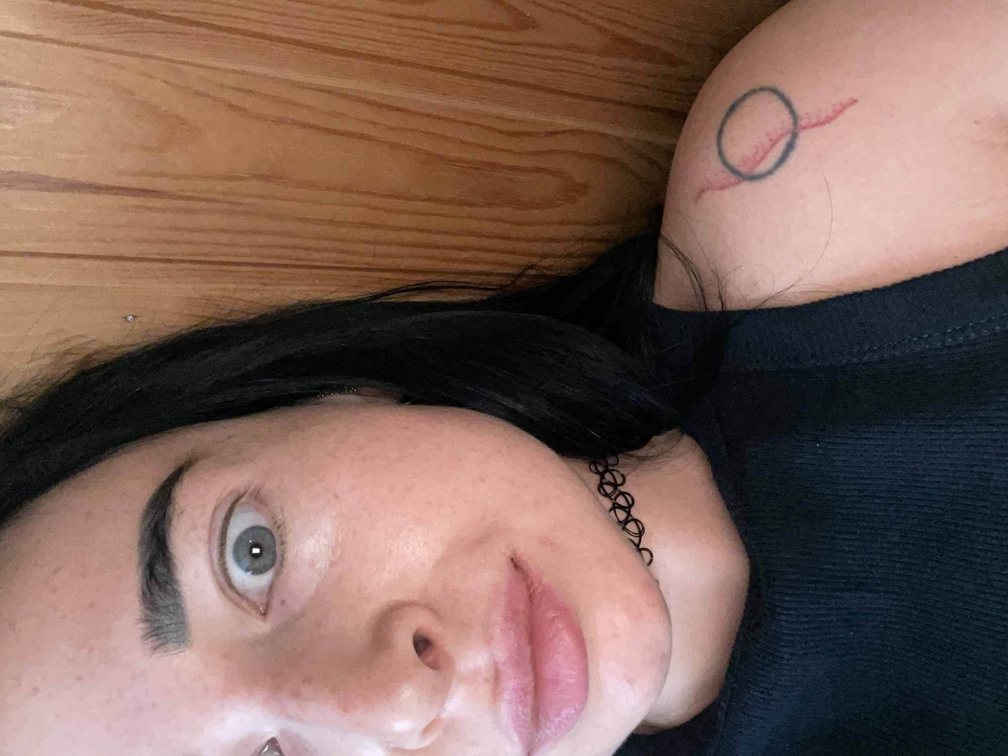The image shows a close-up of a young woman's face and shoulder, taken from a slightly elevated angle. She has fair skin with freckles, dark eyebrows, and light blue-grey eyes. Her lips are full and appear to have a pink lipstick on. She is wearing a black choker necklace and a black sleeveless top. On her left shoulder, she has a tattoo of a circle with a line through it, with some red coloring within the circle. The background is a wooden surface with vertical planks. The lighting appears to be natural, coming from the right side of the frame, creating soft shadows. The woman's expression is neutral, with her eyes wide open.