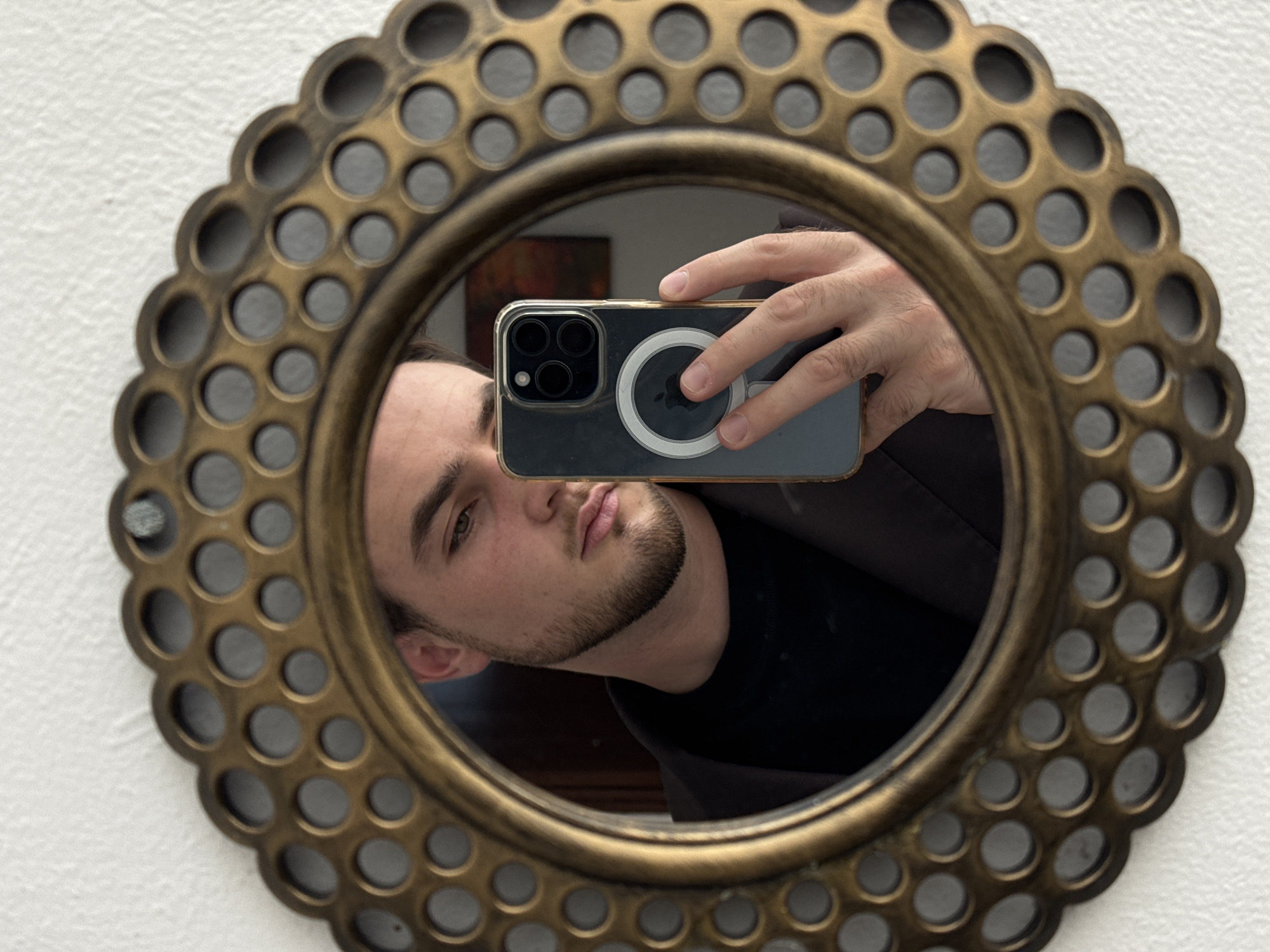 A mirror selfie captured indoors is the main subject of the image. The mirror, which is round, has an intricate bronze or gold-toned frame with a repeating pattern of smaller circles cut out around its perimeter. The mirror is mounted on a white wall.
Reflected in the mirror is a man taking the selfie. He is holding a smartphone with a clear case, showcasing a MagSafe charger attached to the back. The phone's camera array is visible, indicating it may be an iPhone model. The man has dark hair, fair skin, and a neatly trimmed beard. He is wearing a black shirt or top.
The background visible through the mirror is somewhat blurred, but hints at interior furniture and decor. No information about the location of San Bernardino Tlaxcalancingo, Mexico is discernable from the image.