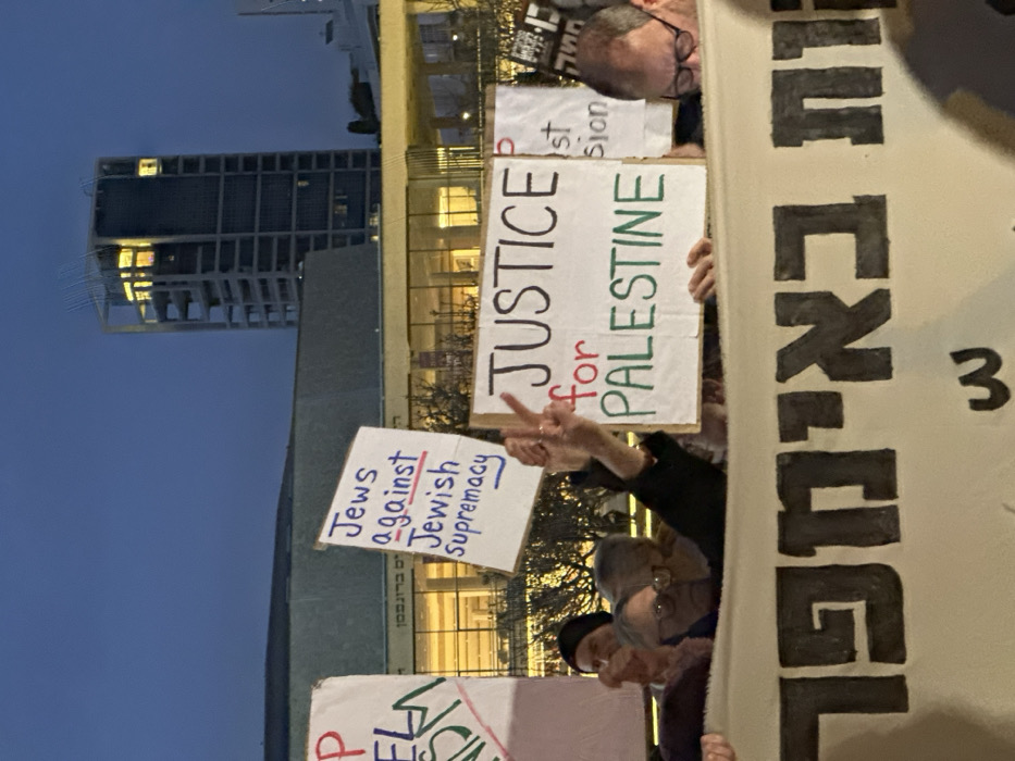 The image depicts a nighttime public demonstration in central Tel Aviv, Israel, specifically near the Tel Aviv Performing Arts Center. The background features the illuminated glass facade of the Performing Arts Center, with the Hebrew text "הישראלי ברונפמן" visible on its structure. Behind it, a tall, multi-storied modern building or skyscraper is partially visible against a dark blue sky.
In the foreground, several individuals, appearing to be adults, are participating in the demonstration. One person on the left wears glasses. A hand in the center raises a V-sign (peace/victory gesture). A man with glasses is partially visible on the right.
The individuals are holding various signs and a large banner. A prominent white sign in the center displays "JUSTICE for PALESTINE" with "for" in red and "PALESTINE" in green. To its left, another white sign reads "Jews against Jewish supremacy" in blue text, with "against" underlined in red. A large, light-colored banner with dark Hebrew letters, partially reading "ות באימפר", spans the lower section of the frame, held by demonstrators. The scene is illuminated by artificial lights from the city and background buildings.