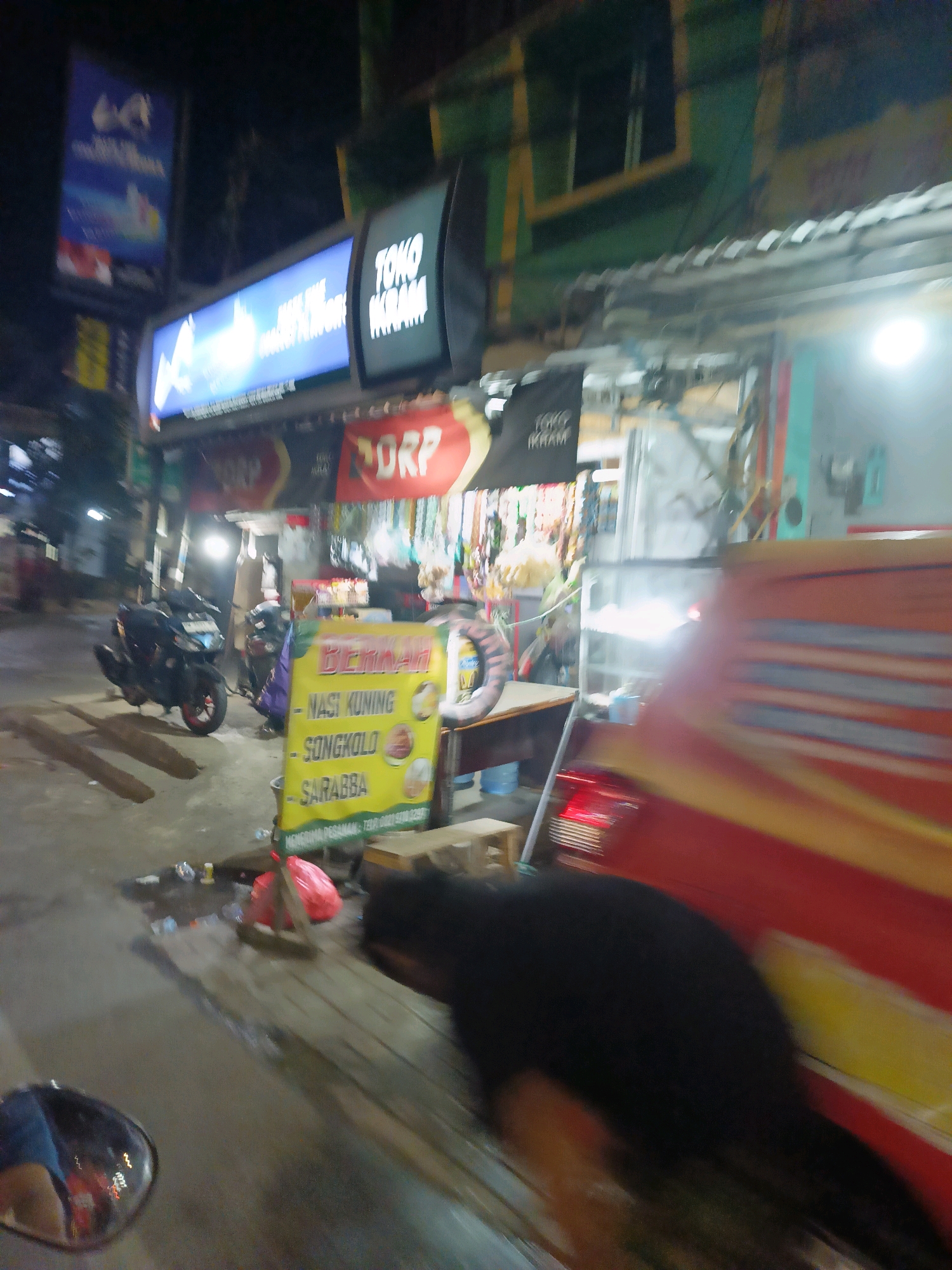 The image depicts a street scene at night in Makassar, Indonesia. A blurry red and yellow vehicle dominates the right side of the frame, partially obscuring a storefront. To the left, a motorcycle is parked on the street. In the foreground, a yellow sign displays text in Indonesian, including "NASI KUNING," "SONGKOLO," and "SARABBA," indicating food items offered. Behind the sign, several items are scattered on the ground, including a red plastic bag, bottles, and debris. Above the storefront, illuminated signs are visible, one in blue with white text and another black sign with white lettering that reads "TOKO IKRAM." The overall impression is of a busy, bustling street with food vendors and shops.