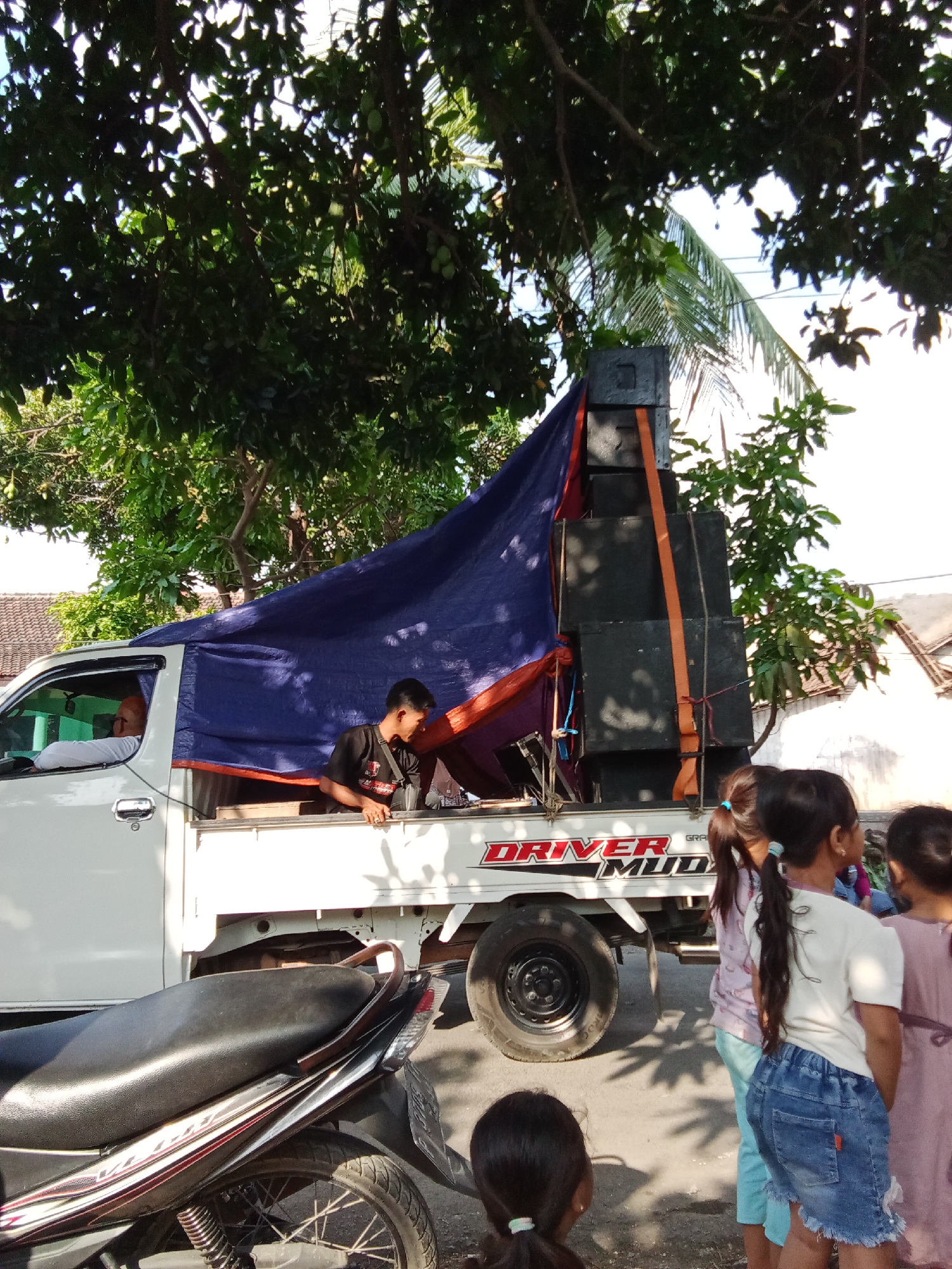In Trebungan, Indonesia, a white pickup truck is parked on a street. The truck bed is covered with a blue tarp, with some of the tarp hanging over the side. Stacked on the truck bed are several black speaker cabinets, secured with orange straps. A young man in a black t-shirt is visible leaning over the speakers, appearing to be working on them. In the driver's side of the truck cab, a person wearing a white shirt and an orange headscarf is seated. To the right of the truck, three young girls are standing. The girl in the foreground is wearing a white t-shirt and denim shorts. Another girl to her left wears a pink patterned shirt and light blue pants. The third girl on the far right is wearing a pink dress. A motorcycle is parked in front of the truck, partially visible on the left side of the frame. Trees with green foliage and some visible fruit are casting shadows on the scene, suggesting it is daytime. Buildings with tiled roofs are visible in the background.