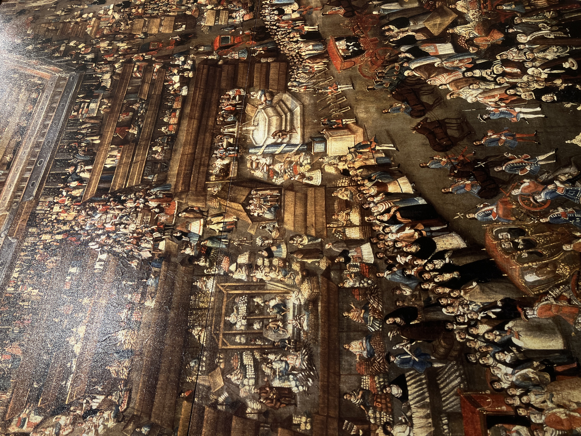 This historical painting, likely from the colonial era in Mexico City, depicts a grand and densely populated public event viewed from a high vantage point. The scene is bustling with hundreds of people spread across streets, squares, and architectural structures.
The subjects include a multitude of people from various social strata, all dressed in detailed period attire. Many figures are seen in military-style uniforms with blue and red coats, some carrying spears or muskets, suggesting a procession or parade. Others are dressed in darker clerical or academic robes. Women are depicted in a variety of dresses and elaborate headwear. Large crowds line buildings with balconies and fill the public spaces, observing the event. Several ornate, horse-drawn carriages, notably a few in red and gold, are prominent, indicating the movement of important figures. Figures on horseback are also visible among the throngs.
A multi-tiered stone fountain serves as a central object in one of the public squares. The setting features detailed colonial architecture with columns, arched passageways, and multi-story buildings. On the left side, structures resembling market stalls or temporary viewing stands are packed with spectators.
The activity appears to be a significant public spectacle, possibly a royal entry, a religious procession, or a major civic celebration. The atmosphere suggests order and a sense of occasion. The bright, even lighting indicates the event is taking place during the daytime. No specific text is visible within the painting itself. The overall image displays surface texture or glare, suggesting it is a photograph of a painted artwork.
