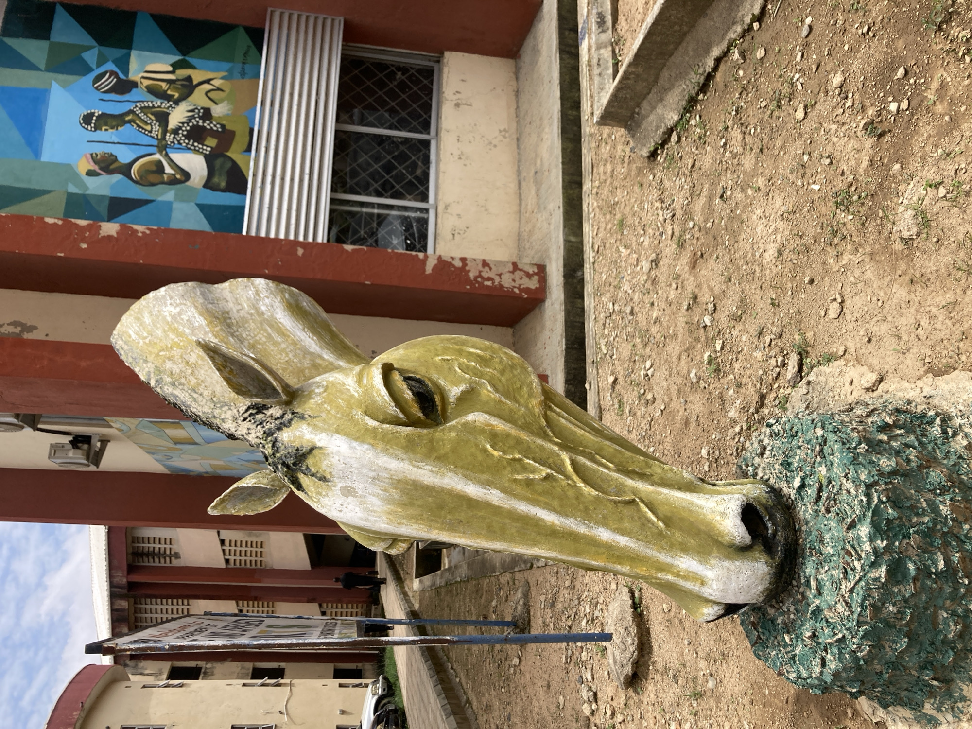 A close-up shot focuses on the head of a weathered, light green horse sculpture, its surface showing peeling paint and black grime. The sculpture is mounted on a short, cylindrical pedestal covered in textured green paint. In the background, a red and cream colored building is visible, adorned with a vibrant mural depicting three individuals in traditional attire. To the left of the sculpture, a metal signpost with blue poles stands on a concrete base, with a partially legible sign. A person in black clothing walks away from the camera in the middle ground, near the building entrance. The ground is covered in loose dirt and small stones. The scene appears to be an outdoor public space, possibly a university campus, given the building's architecture and the presence of signage. The image quality is good, with natural lighting and clear details.