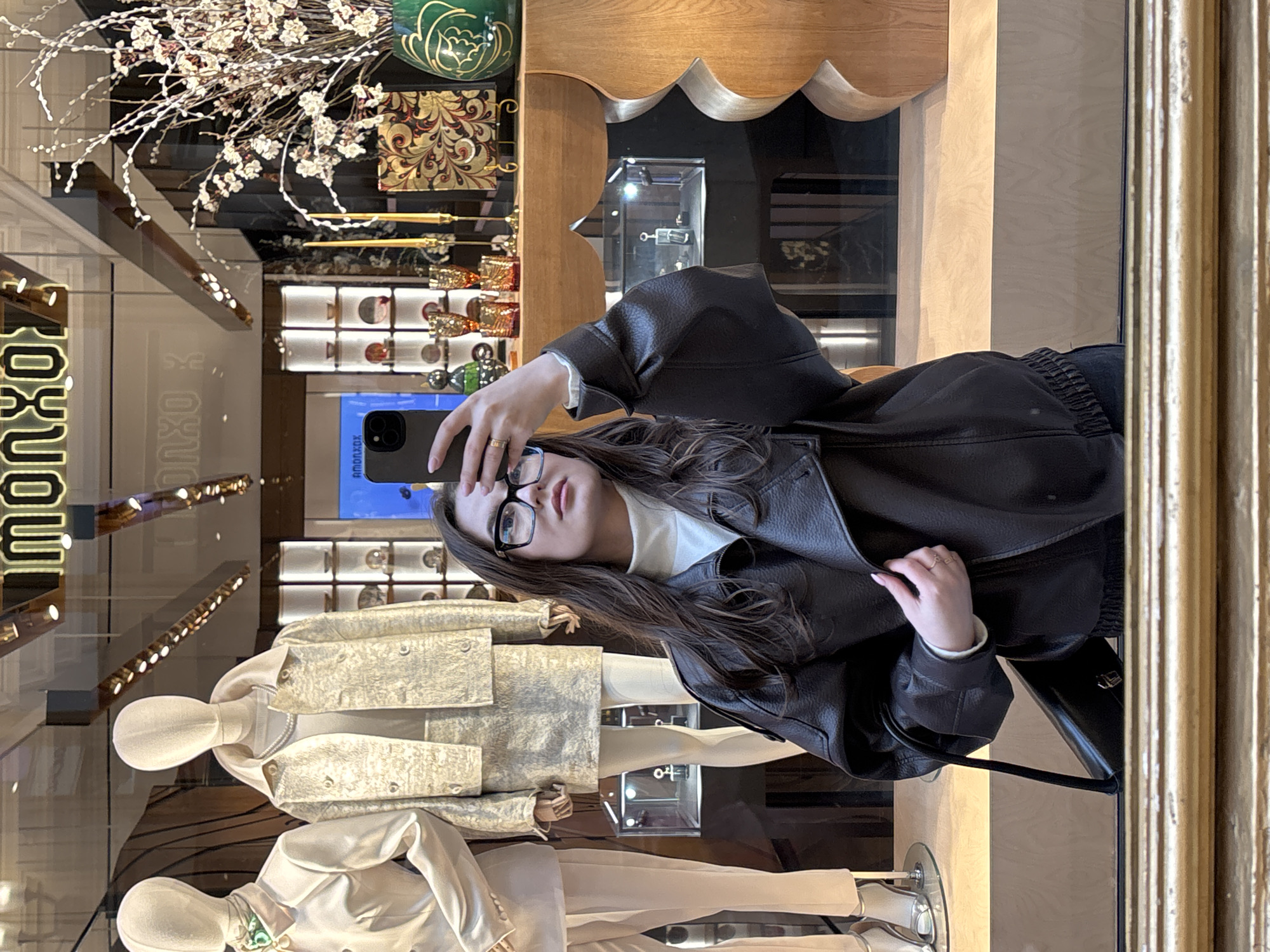 The image shows a woman taking a selfie in front of a shop window. The shop appears to be a luxury goods store, with mannequins displaying high-end clothing. The woman is wearing a black leather jacket, glasses, and a white top. The reflection in the window shows the interior of the store, with display cases and decorative items. The sign "TSUM" is partially visible in the window, indicating that the location is likely the TSUM department store in Moscow, Russia.