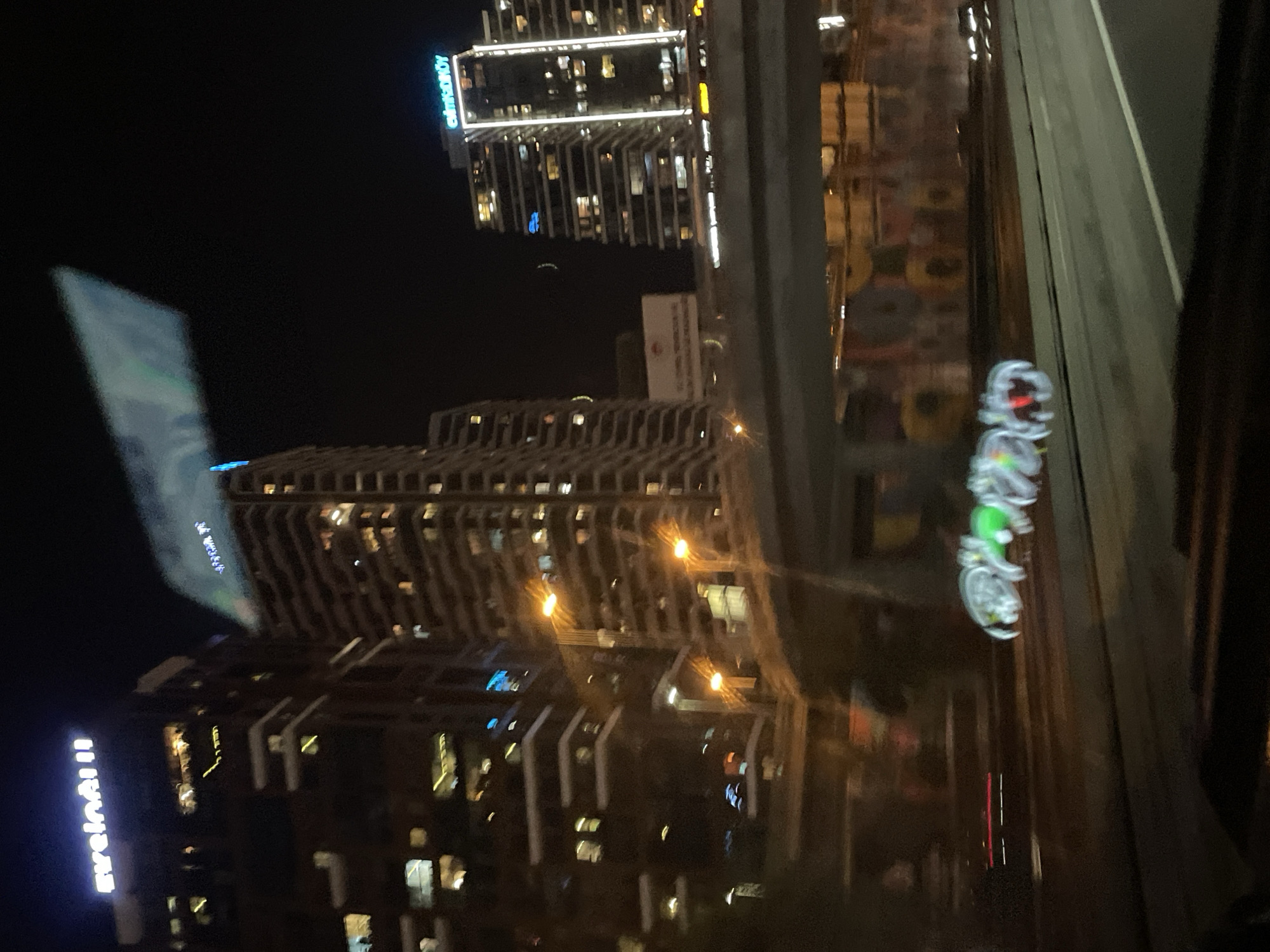 The image depicts a nighttime scene in Kadıköy, Turkey. In the foreground, a blurred light trail, possibly from a moving vehicle's decorative lights, forms abstract shapes in shades of green and red. The background features illuminated high-rise buildings with visible windows, suggesting an urban environment. One building prominently displays the word "Evelinall" in white neon letters. Another building has "ezimKADIKÖY" in blue neon letters. A highway overpass stretches across the mid-ground, with the underside of the overpass adorned with colorful graffiti art. The sky is dark, indicating it is night. The overall impression is that of a city view from a moving vehicle at night, with a focus on illuminated architecture and light trails.