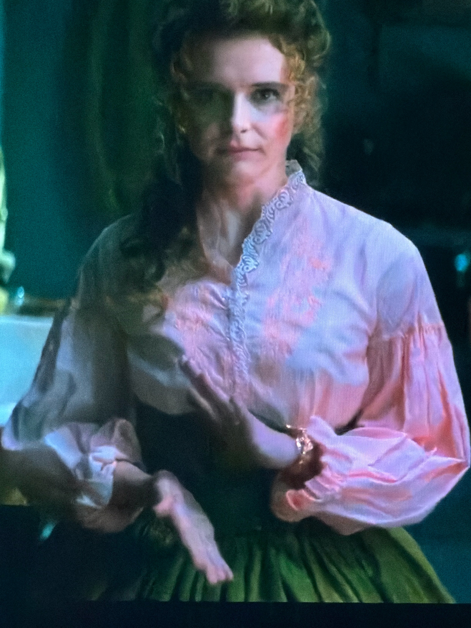 In Agachaul, Russia, a woman with curly brown hair and smudged red makeup on her cheeks is shown from the chest up. She is wearing a pink blouse with puffed sleeves and intricate white lace trim around the neckline. Her hands are clasped over her chest, with one hand adorned with a ring. A green skirt is visible at the bottom of the frame. The background is dark and out of focus, with hints of green and blue light.