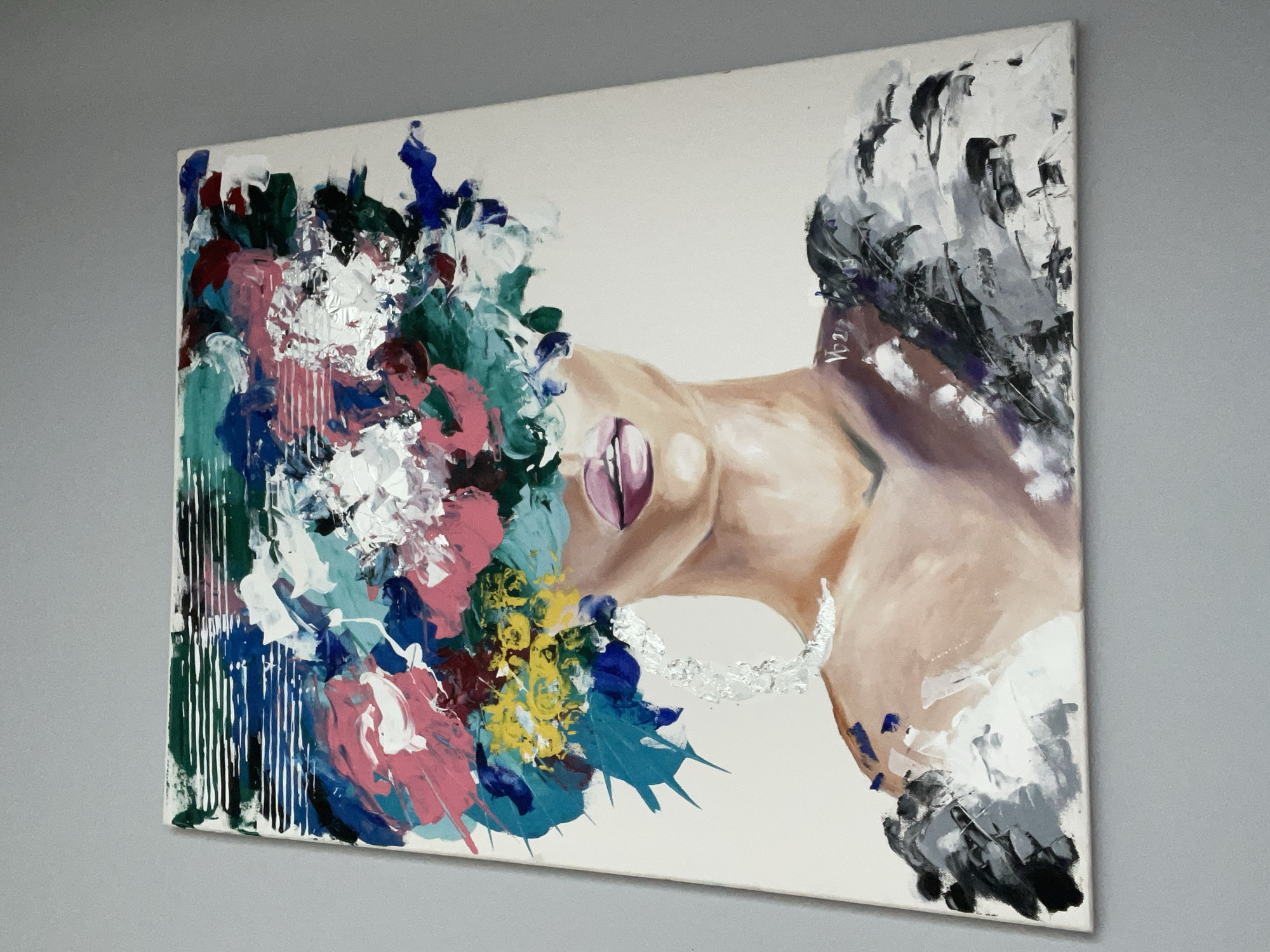 A painting on a white canvas depicts a partial profile of a human figure emerging from a mass of vibrant colors and abstract strokes. The painting is hung against a light gray wall. The human figure's skin tone is rendered in shades of beige and brown, with the lips painted in a mauve or purple hue. The neck is slightly arched, and there is a hint of a collarbone visible. Silver leaf accents are applied to the throat and chest area. The figure is almost entirely obscured by an explosion of colors on the left side of the canvas. These colors include pink, white, blue, green, and yellow, applied with thick, expressive strokes. Some areas of the colors drip vertically. On the right side of the canvas, the figure is partially obscured by an array of black and gray brushstrokes. The painting is likely an expressionist or abstract work, focusing more on emotion and form rather than precise representation. The location is undeterminable as there is no identifiable information about Sofia, Bulgaria in the image.