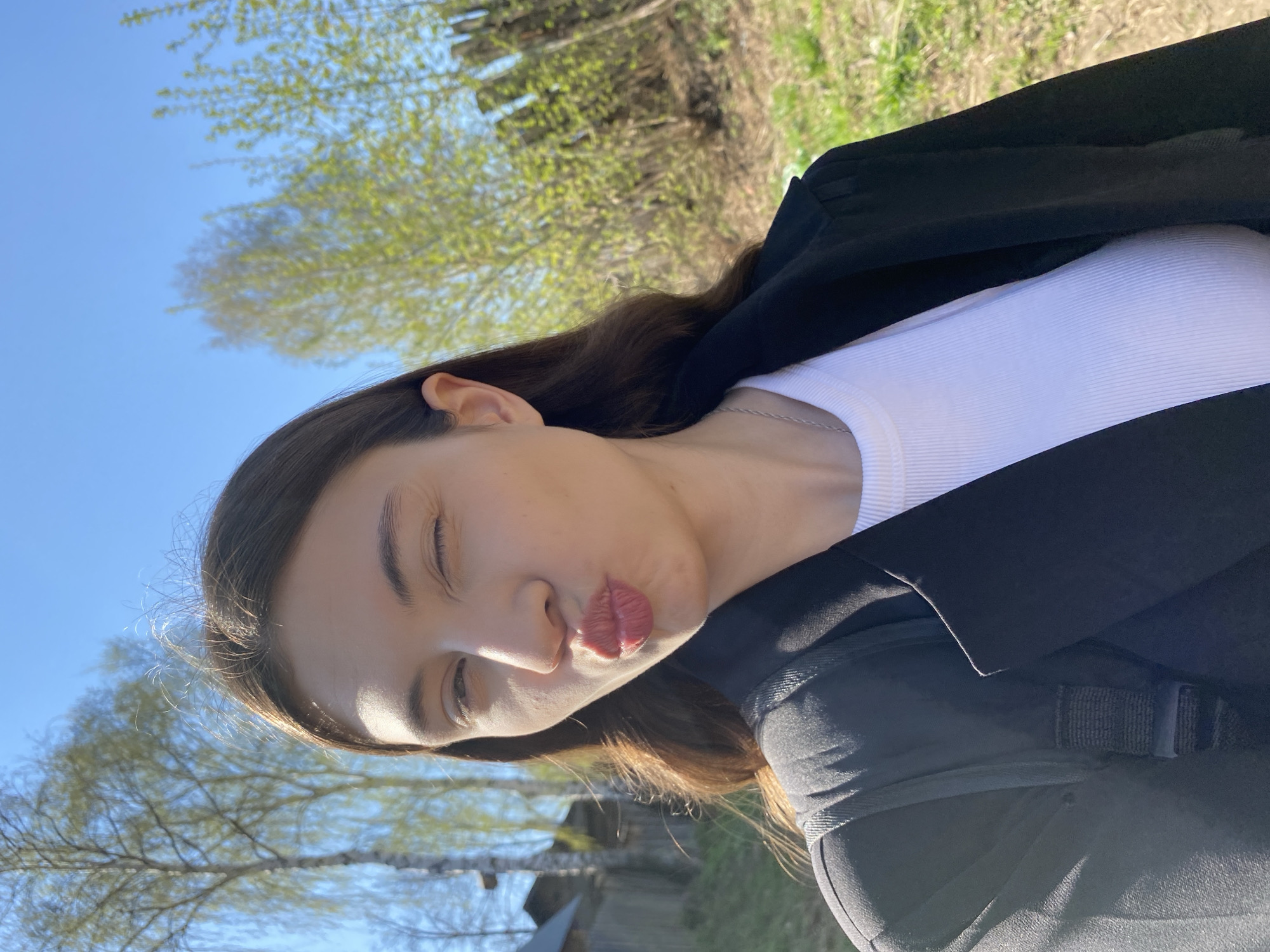 A young woman is posing for a selfie outdoors. She is wearing a white ribbed top and a black blazer or jacket. Her long brown hair falls over her shoulder. Her eyes are partially closed, and she is puckering her lips in a kissing gesture.
The background includes trees with budding green leaves and a clear blue sky. The location is likely a park or green area. It is impossible to independently verify the specific location in Votkinsk, Russia, from the image alone.