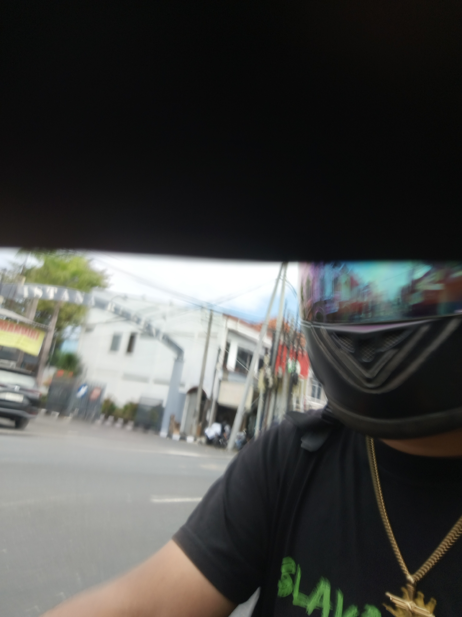 The image shows a first-person perspective of someone riding a motorcycle in Denpasar, Indonesia. The rider is wearing a black t-shirt and a black helmet with a reflective visor. A gold chain with a pendant is visible on the t-shirt. The background is blurred due to motion and shows buildings, trees, and a street with other vehicles. A sign with text, partially visible as "ISTANA," is also present in the background, suggesting a commercial or residential area. The overall scene depicts everyday street life in the city.