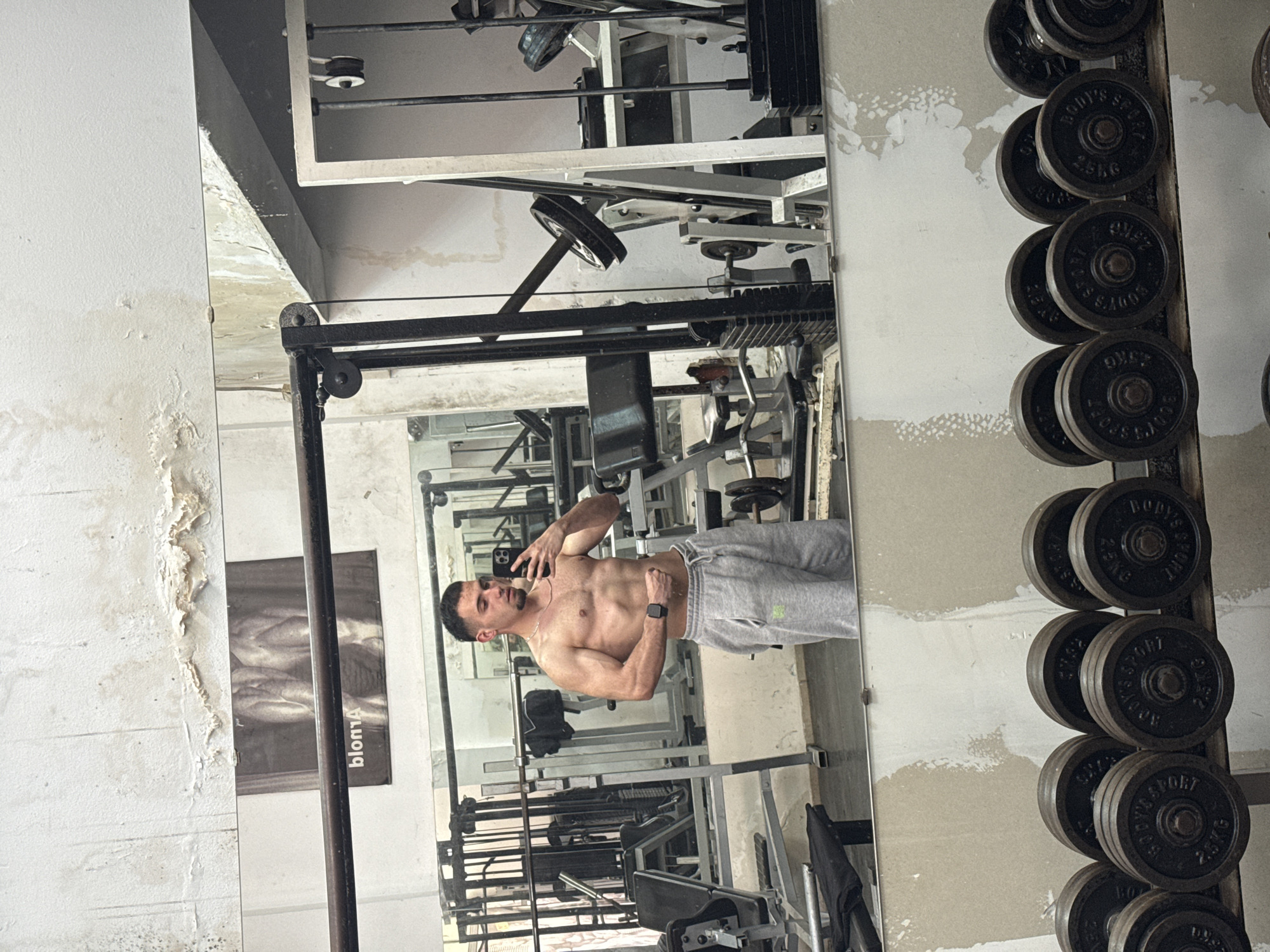 A shirtless man with a lean, muscular physique is captured in a mirror selfie within what appears to be a gym. He is wearing light grey sweatpants and a smartwatch on his left wrist, holding a black smartphone with his right hand to take the photo. His left hand is placed on his waist, and he is posing to showcase his abdominal and arm muscles.
The mirror reflects various gym equipment, including weight machines with cables and pulleys, and a black weight rack on the right side of the image, stacked with circular weight plates. Some plates are labeled "BODY'S SPORT" and indicate "2.5 KG". A black object, possibly a gym bag or clothing, is draped over some equipment in the reflection.
The walls visible in the foreground and reflection appear discolored and worn, with sections of peeling paint or plaster, suggesting an older facility or a distressed aesthetic. A poster or chart with human anatomy, partially visible as "blom4" (likely a reflection of "body anatomy" or similar) is visible behind the man's reflection. The overall image orientation is rotated 90 degrees counter-clockwise, making the man and his reflection appear upright relative to the frame, but the gym elements and walls are on their side. The scene is indoors, in Shumen, Bulgaria. The man appears confident while posing.