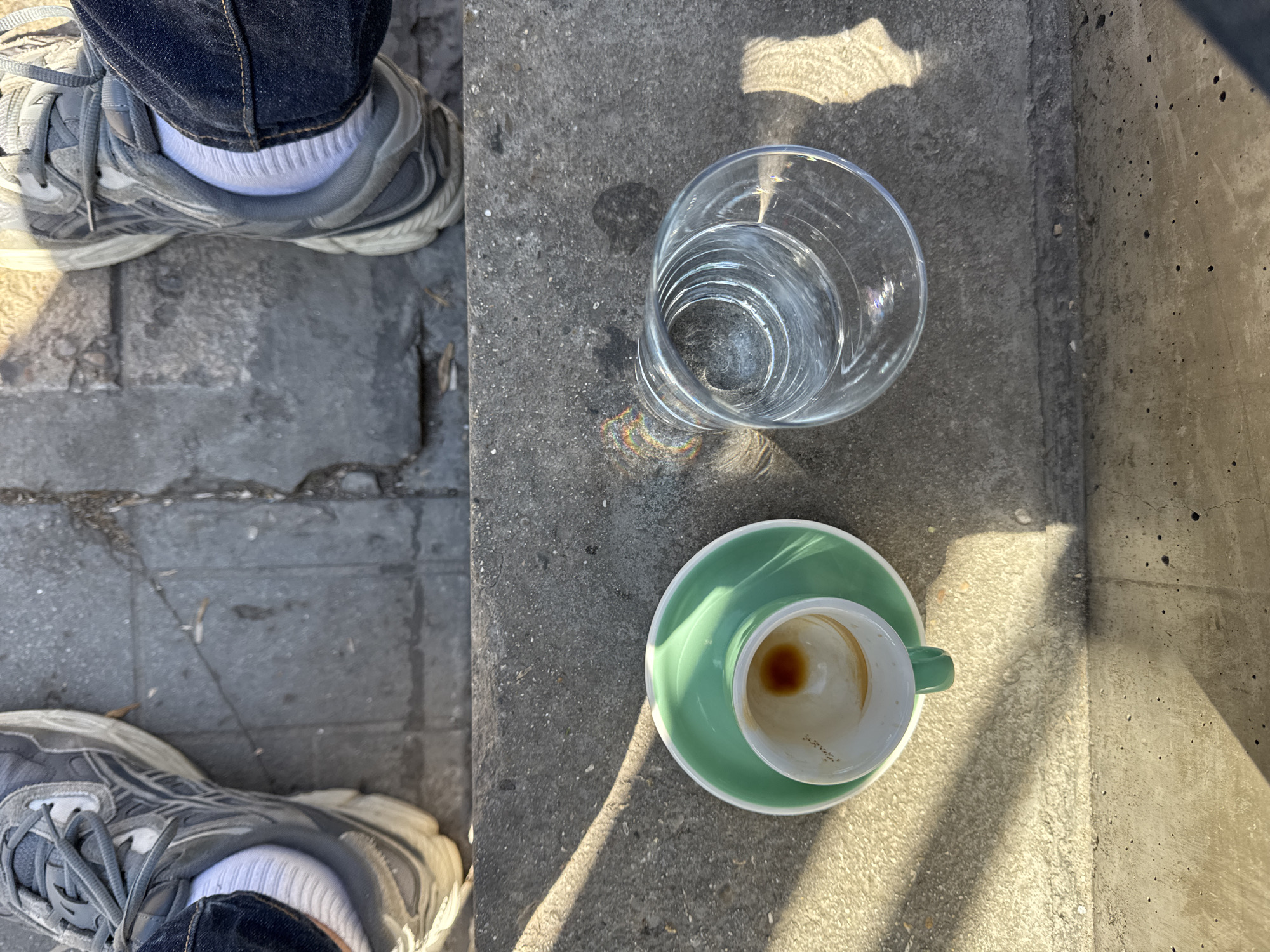 The scene is an outdoor setting, possibly a cafe or patio in Sofia, Bulgaria. The ground is a mix of stone pavers and concrete, partially in sunlight. A person's legs, clad in blue jeans, white socks, and gray athletic shoes, are visible in the left of the frame. On the concrete surface, a clear glass of water sits near a used coffee cup and saucer, the cup is pale green with a small amount of dark liquid residue inside.