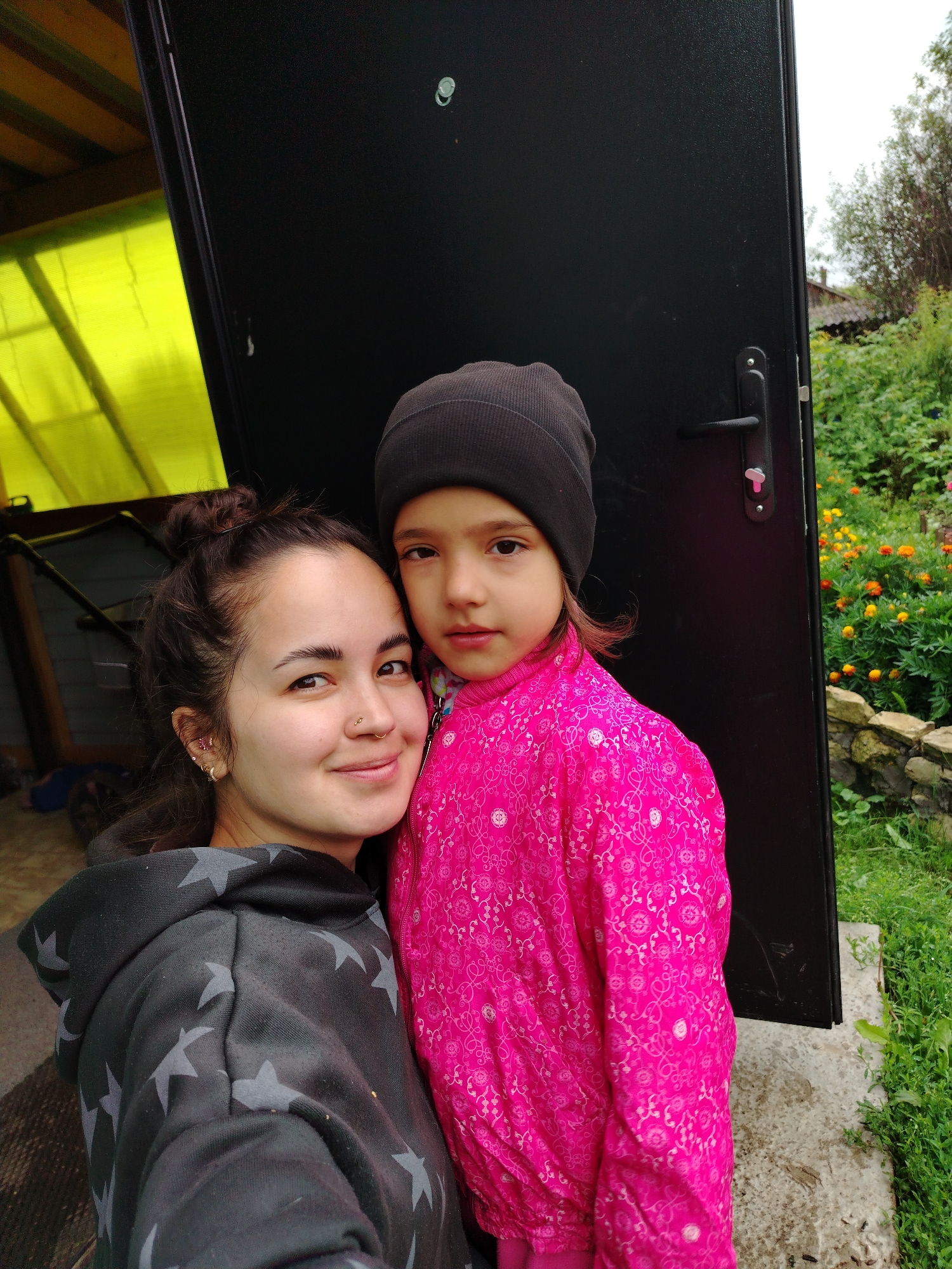 In Гожан, Russia, two people are pictured standing outdoors, partially framed by a large, black door. The person on the left, likely an adult, is wearing a dark grey hooded sweatshirt with a pattern of light grey stars. Their hair is pulled up into a bun, and they are smiling, looking at the camera. They have earrings and a nose ring. To their right, a young person, likely a child, is wearing a bright pink jacket with a patterned design and a dark grey beanie. The child is looking forward, with a neutral expression. Behind them, the black door is slightly ajar, revealing a glimpse of a yellow structure on the left and a garden with flowers and greenery on the right. The ground appears to be a mix of concrete and dirt.