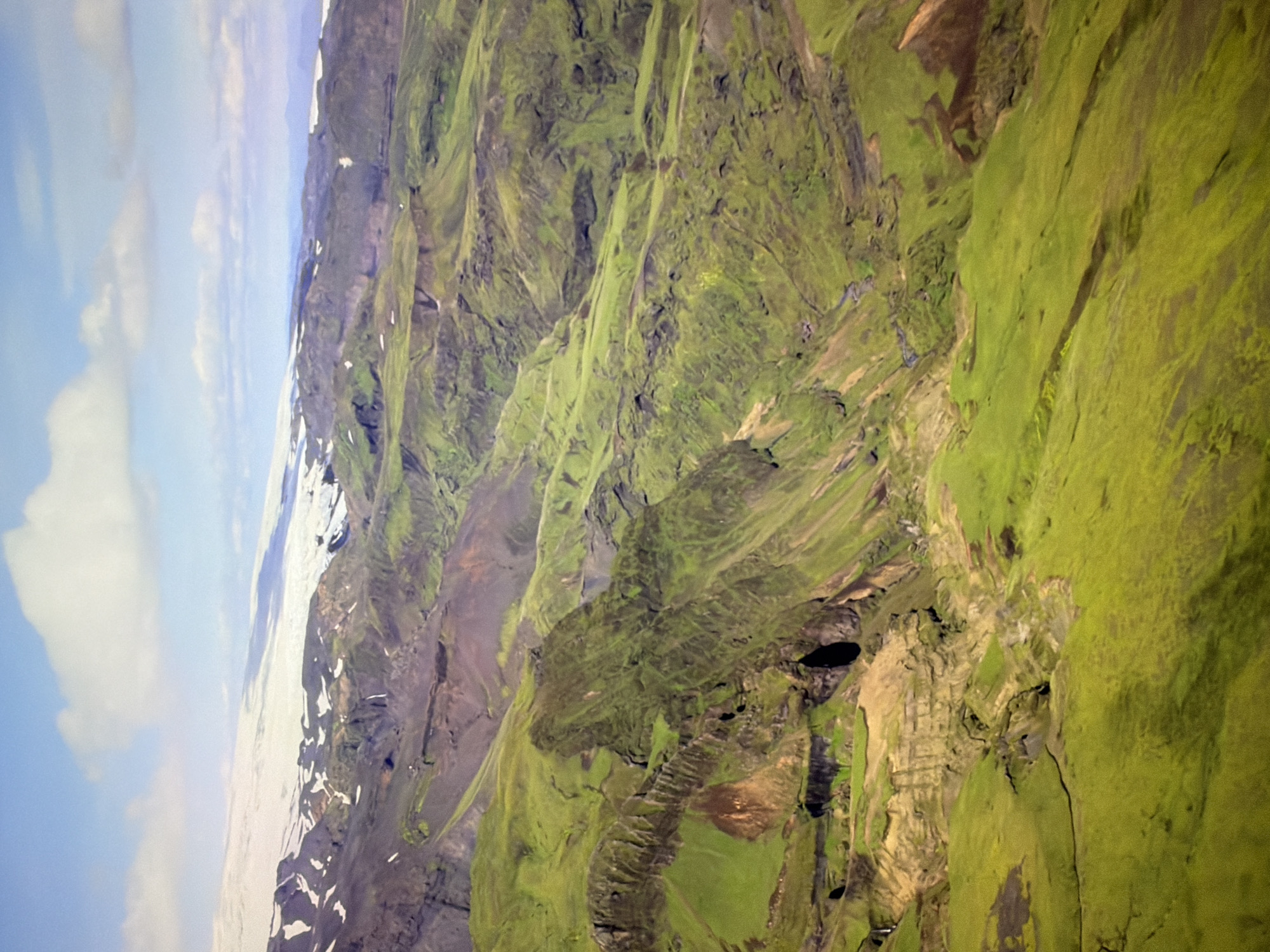 The image shows an aerial view of a rugged, volcanic landscape covered in vibrant green vegetation. In the background, a snow-capped mountain range stretches across the horizon under a partially cloudy blue sky. The terrain is characterized by steep slopes, deep ravines, and exposed rock faces. There are no people, structures, or any signs of urban development visible in the image. The scene is entirely natural and appears to be a remote, undeveloped area. There is no identifiable connection to the city of Kyiv, Ukraine.