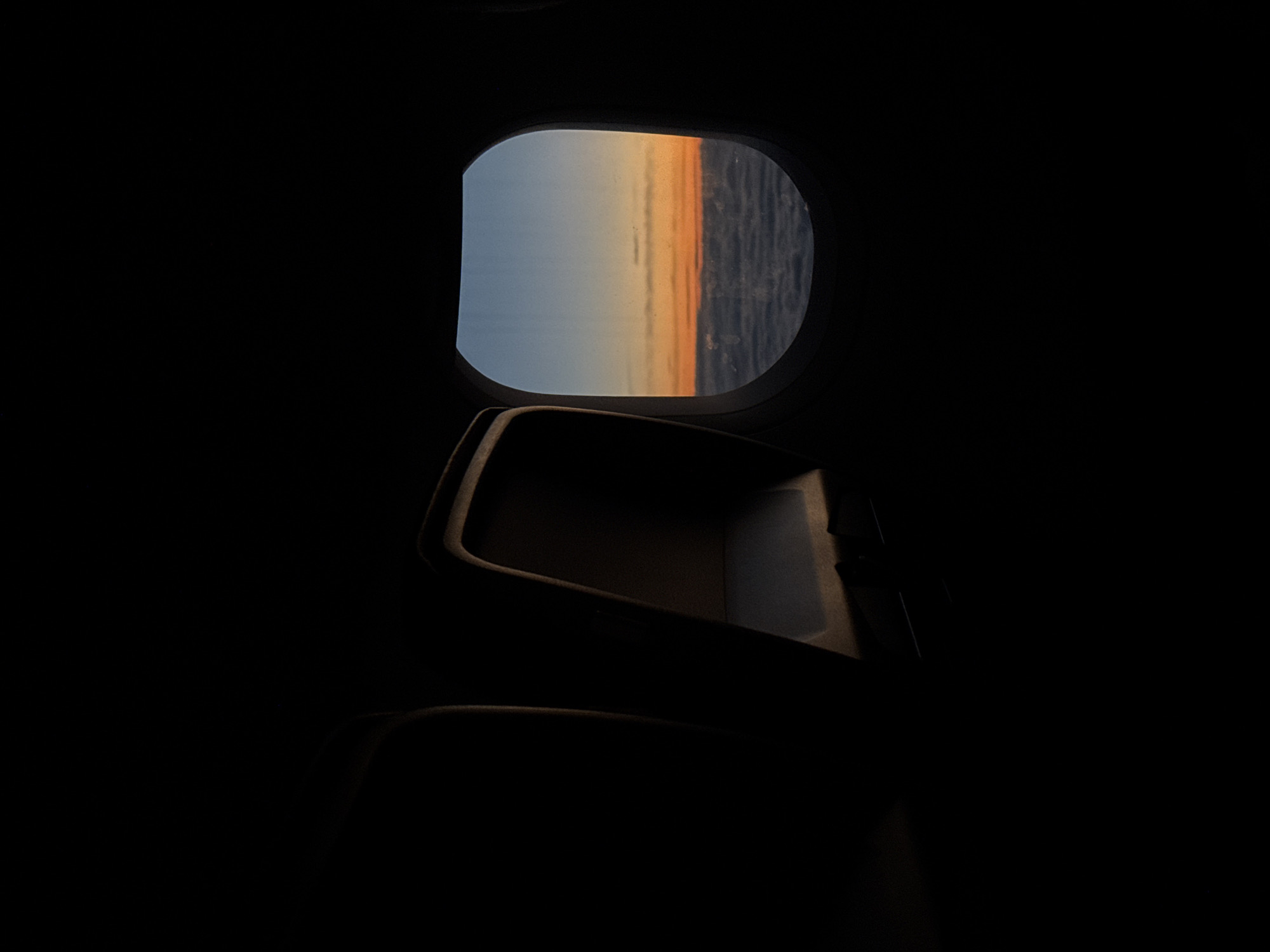 The image depicts the interior of an airplane cabin, specifically the seat and window area. The cabin is dimly lit, with most of the scene obscured by darkness. The only source of illumination is the sunlight streaming in through the window. The window reveals a view of the sky and the ocean or horizon. The sky exhibits a gradient of color, transitioning from blue to orange. The image provides no information about Dortmund, Germany.