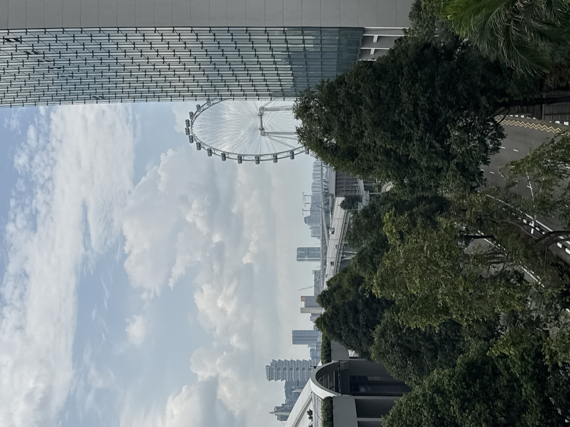 The image depicts an aerial view of a cityscape in Singapore, characterized by modern architecture and lush greenery. In the foreground, a cluster of mature trees with dense, dark green foliage dominates the lower portion of the frame, with a road running alongside them. To the left, a portion of a building with a curved, gray roofline is visible.
The mid-ground reveals a wider expanse of trees, and beyond them, a multi-lane road or highway with vehicles can be seen. In the background, a large Ferris wheel, likely the Singapore Flyer, stands prominently, its white structure contrasting with the sky. Several skyscrapers and other tall buildings form the city's skyline, some with distinctive architectural designs. The sky is partly cloudy, with patches of blue visible between the white cumulus clouds. The overall scene suggests a view from an elevated vantage point, overlooking a blend of urban development and natural elements within Singapore.