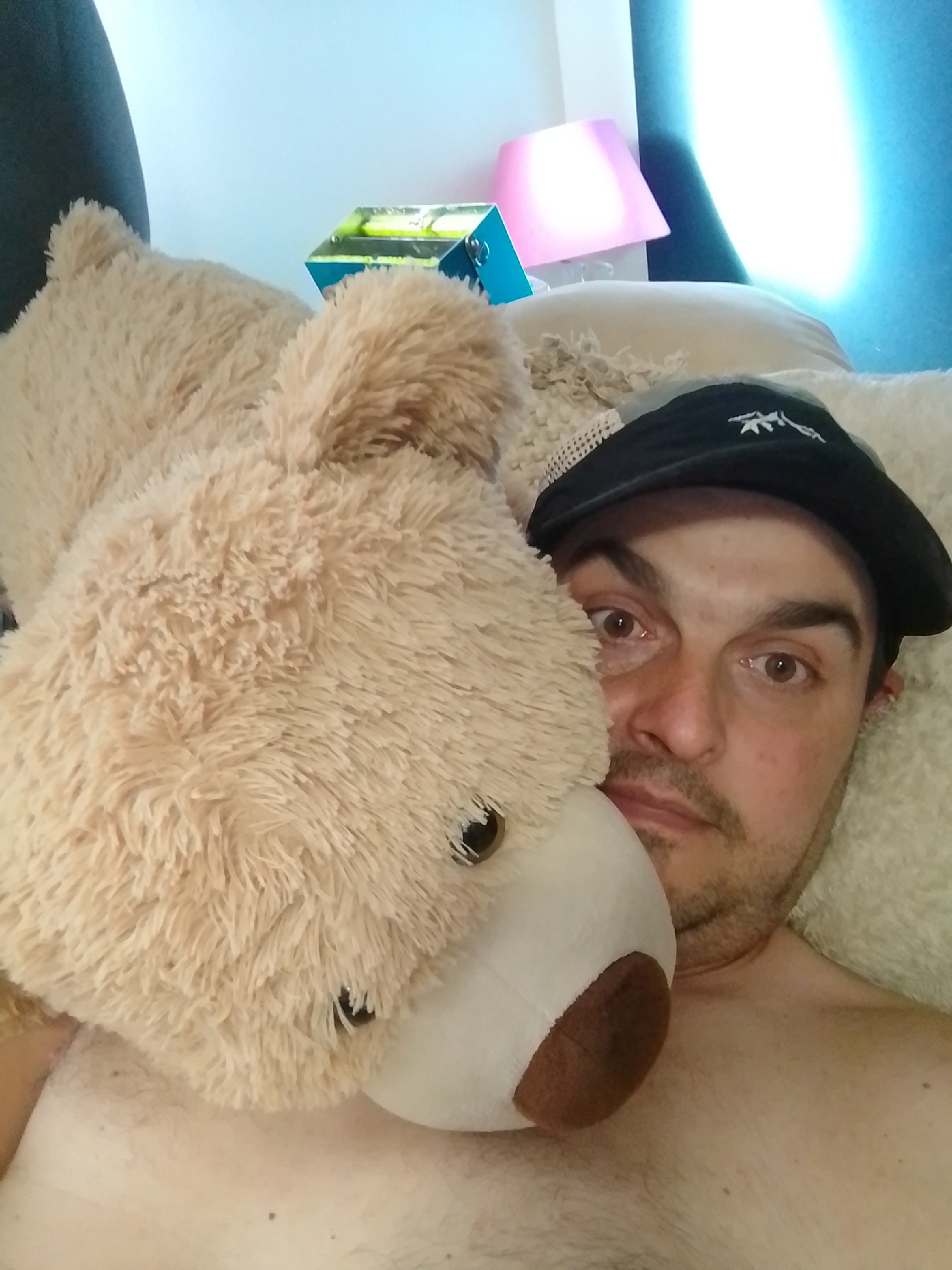A man wearing a black baseball cap is lying in bed, with a large stuffed teddy bear resting on his chest. The man's head is turned towards the camera, and he is looking directly at it with his eyes open. The teddy bear's head is covering part of the man's face, and its nose is resting on the man's cheek. The scene appears to be indoors, likely in a bedroom, with a pink lamp and a blue box visible in the background. The lighting is dim, with a bright light source coming from the upper right corner of the image. The overall impression is one of casual relaxation or perhaps an attempt at a playful selfie.