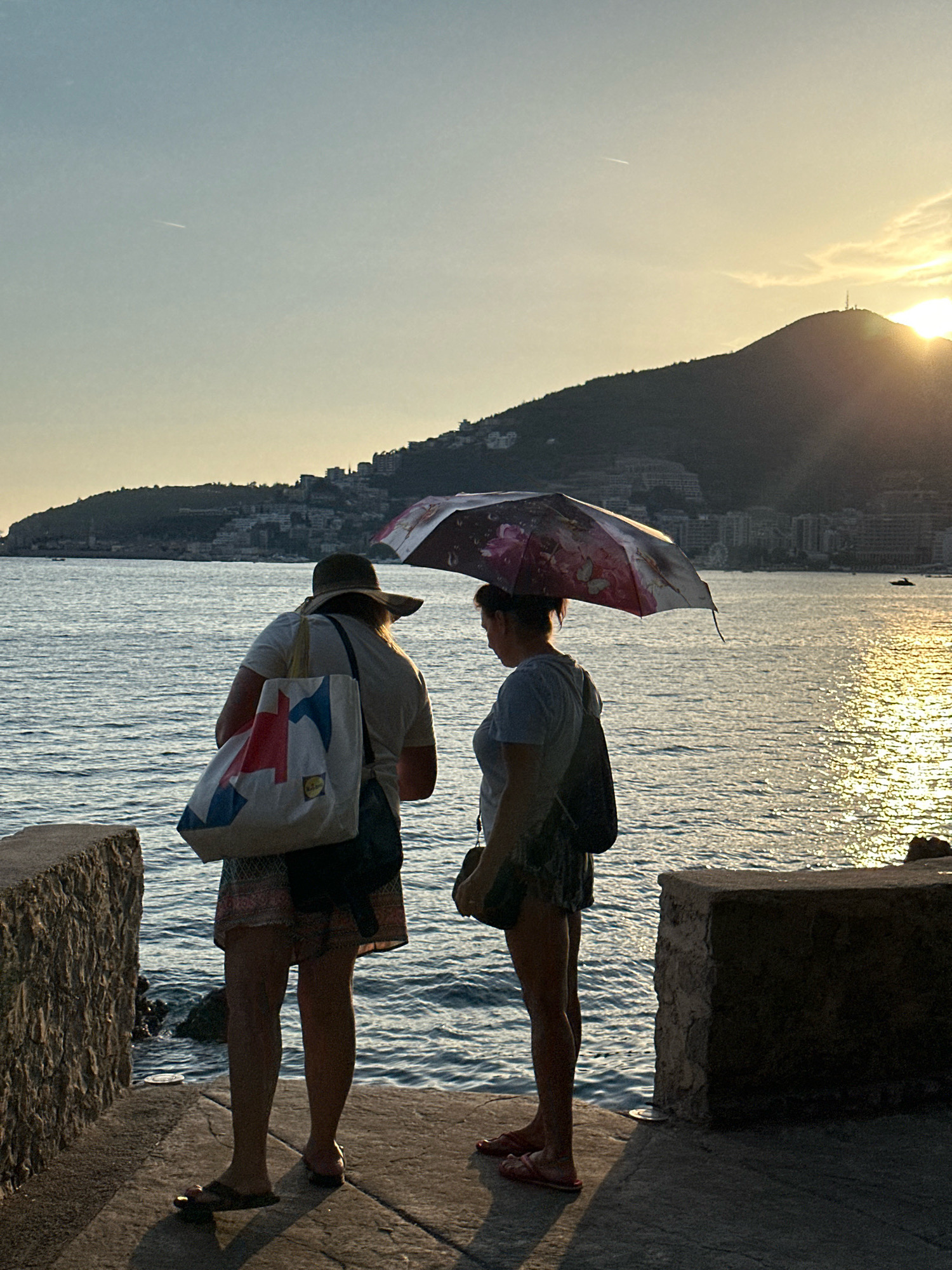 Two women stand on a concrete promenade overlooking the Adriatic Sea in Budva, Montenegro. The woman on the left wears a wide-brimmed hat, a light-colored t-shirt, and patterned shorts, carrying a large tote bag with red, blue, and white geometric patterns. She is wearing dark sandals. The woman on the right holds a patterned umbrella over her head and wears a grey t-shirt and patterned shorts, with flip-flops on her feet. Both women are facing away from the viewer, towards the sea and a hillside town in the background. The sun is setting behind a hill on the right side of the frame, casting a warm glow on the water and illuminating a portion of the town. The water is calm with gentle ripples. The promenade is made of rough-hewn stone walls on the left and a lower concrete ledge on the right.