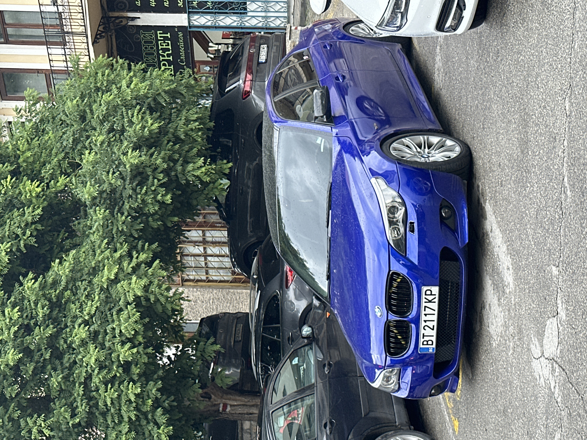 A bright blue BMW sedan is parked on an asphalt surface, in front of other parked cars and a building with a large tree. The BMW's license plate reads "BT 2117 KP". The scene appears to be an urban environment in Veliko Tarnovo, Bulgaria, as indicated by the license plate prefix "BT" which denotes the city of registration. The building behind the cars has multiple windows and balconies, and a sign with Cyrillic text is visible. The ground is cracked and weathered. The lighting suggests it is daytime, possibly overcast. There are no people visible in the image.