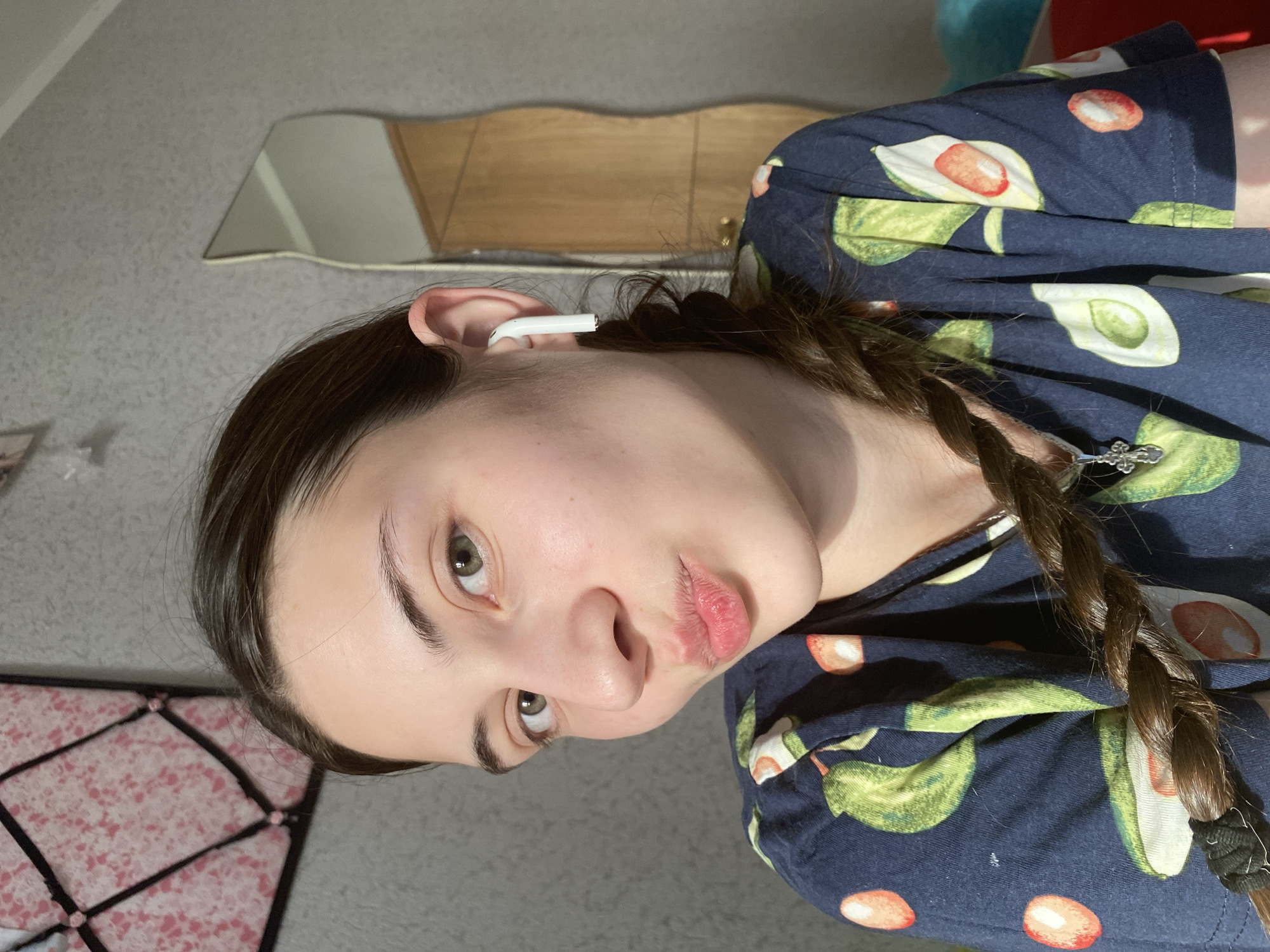 A close-up, tilted shot features a young woman with light skin, dark brown hair styled in a braid, and green eyes. She has an earbud in her ear. She's wearing a dark blue top patterned with avocado images. The background includes a gray carpeted floor and a partially visible mirror. A piece of furniture is reflected in the mirror. A portion of a black and pink object, possibly a chair or cushion, is visible in the lower left corner. It's impossible to confirm the exact location is Votkinsk, Russia based only on the visual elements in the image.
