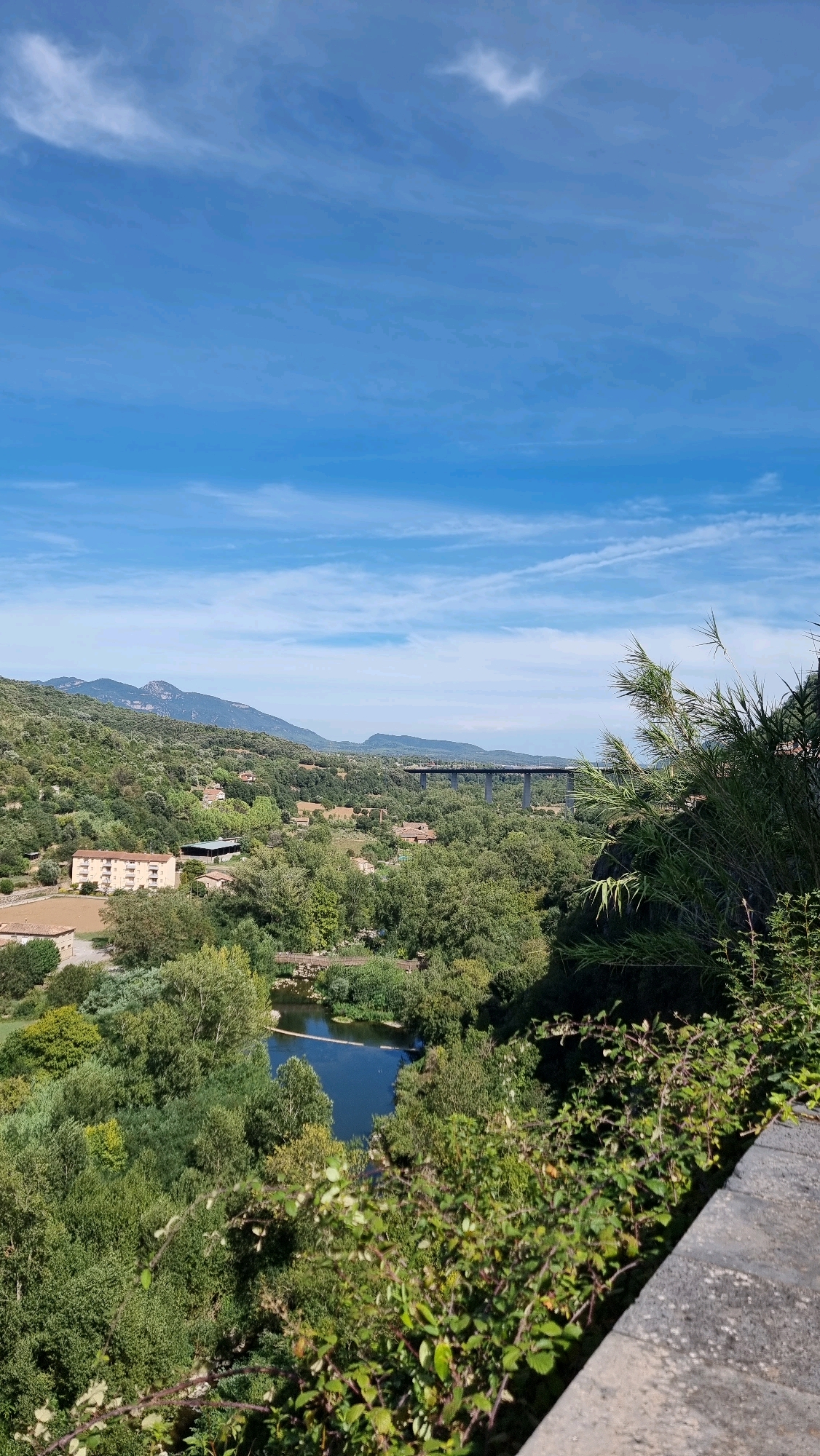This is an eye-level, high-angle, outdoor, full shot photograph taken on a sunny day. The image captures a verdant landscape with a winding river or lake at its center. The water is dark blue and reflects the bright sky. Surrounding the water is a dense forest of lush green trees, with some residential buildings interspersed throughout the foliage. In the mid-ground, a long, dark bridge with multiple piers spans across the landscape in the distance. The background is dominated by rolling hills and mountains under a clear blue sky with wispy white clouds. In the foreground, there is a stone wall or pathway with a dense growth of green bushes and thorny vines cascading over it, partially obscuring the view. The scene appears to be a natural landscape with human infrastructure. The location is identified as Castellfollit De La Roca, Spain. There are no people or animals visible in the image.