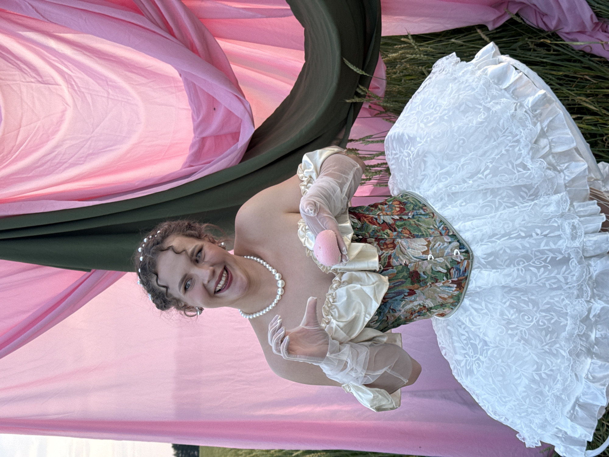 A young woman in a costume resembling a ballet tutu stands in a field of tall grass. She is wearing a white, layered tutu, a floral patterned corset with a light-colored, ruffled bodice, and sheer white opera gloves. A pearl necklace adorns her neck, and a beaded headband is in her curly, brown hair. She is smiling and holding a small, pink object, possibly a macaron or a small soap, in her gloved hands. Behind her, draped fabric creates a backdrop of pink and dark green. The setting appears to be outdoors, with vegetation visible in the foreground and background.