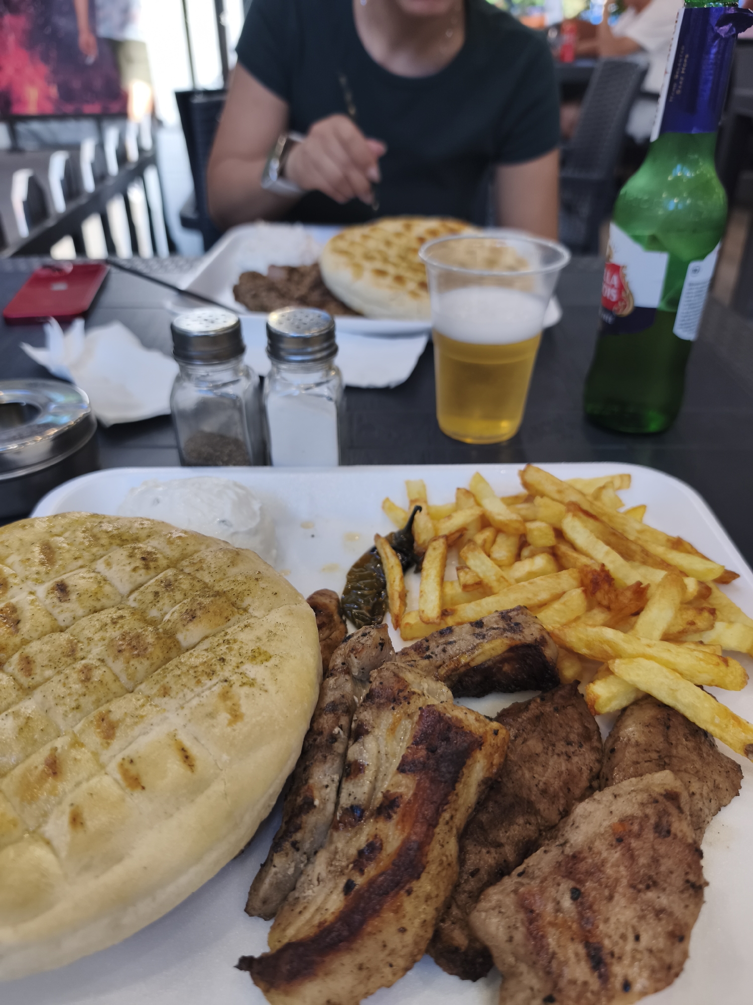 The image shows a close-up of a meal served on a white plate. The meal includes grilled meat, french fries, a flatbread, and a dollop of white sauce, possibly yogurt or sour cream. The grilled meat appears to be various cuts, some with grill marks. The french fries are golden brown. The flatbread is round and appears to be seasoned with herbs.
In the background, a person is visible from the shoulders up, wearing a dark green t-shirt. Their hands are on the table, and they seem to be reaching for something. To the right of the person, a green bottle of Stella Artois beer is partially visible, along with a plastic cup filled with beer. In front of the person, a red smartphone is lying on the table. Salt and pepper shakers are also present on the table.
The setting appears to be an outdoor dining area, possibly a restaurant or cafe, with a glimpse of a blurred background that suggests tables and chairs. The lighting indicates it is daytime. The overall mood of the image is casual, focusing on the food and a relaxed dining experience.