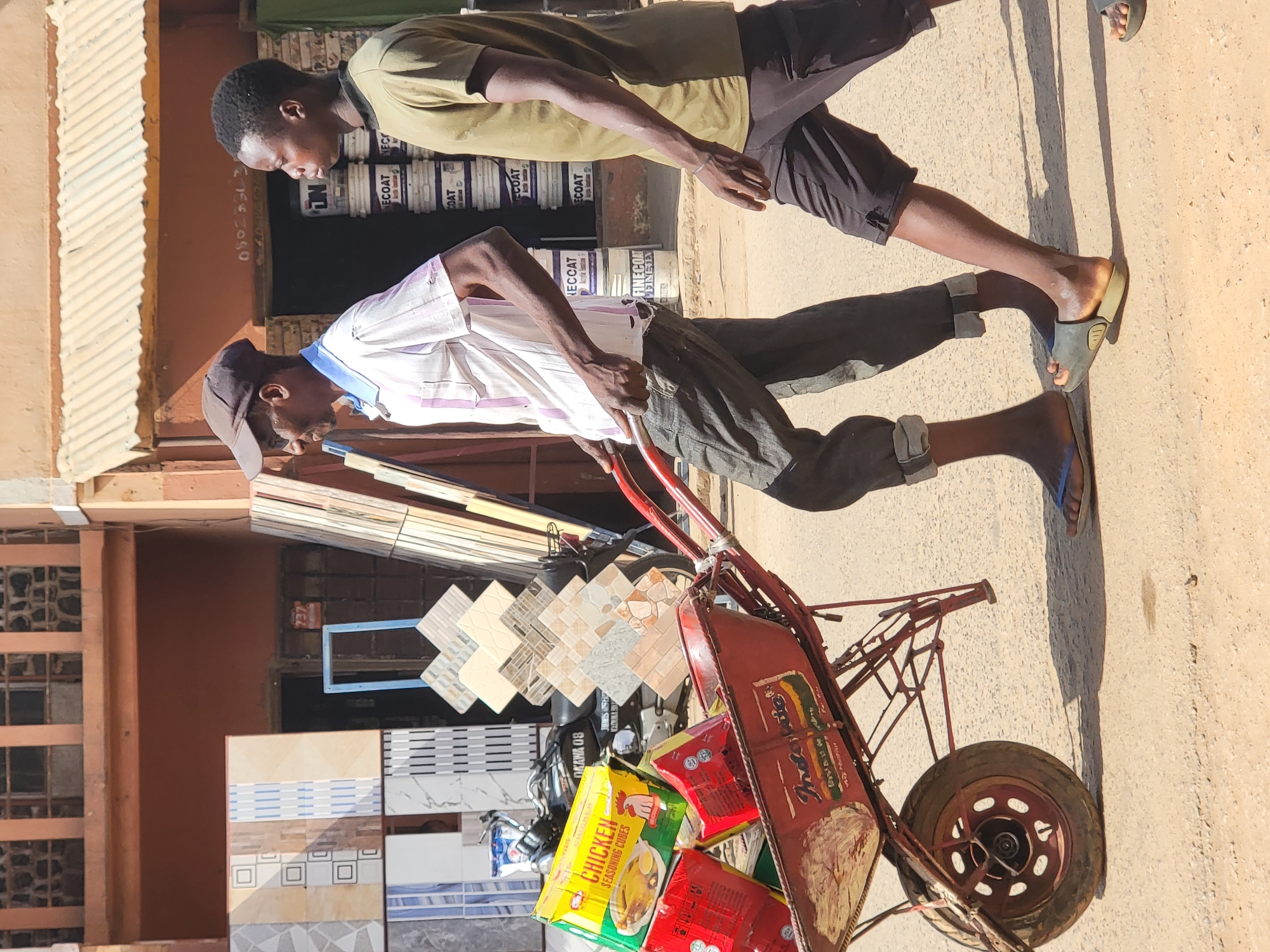 Two dark-skinned men transport goods on a wheelbarrow in the city of Maiburiji, Nigeria. One man in a light, vertically striped shirt and dark pants pushes a red wheelbarrow, filled with boxes of chicken seasoning cubes, red packets, and what appear to be ceramic tiles. The other man wears a green shirt and dark shorts and appears to be helping the first man. In the background are tiles displayed for sale, paint buckets, and a building with barred windows. The environment is urban, likely a commercial area.