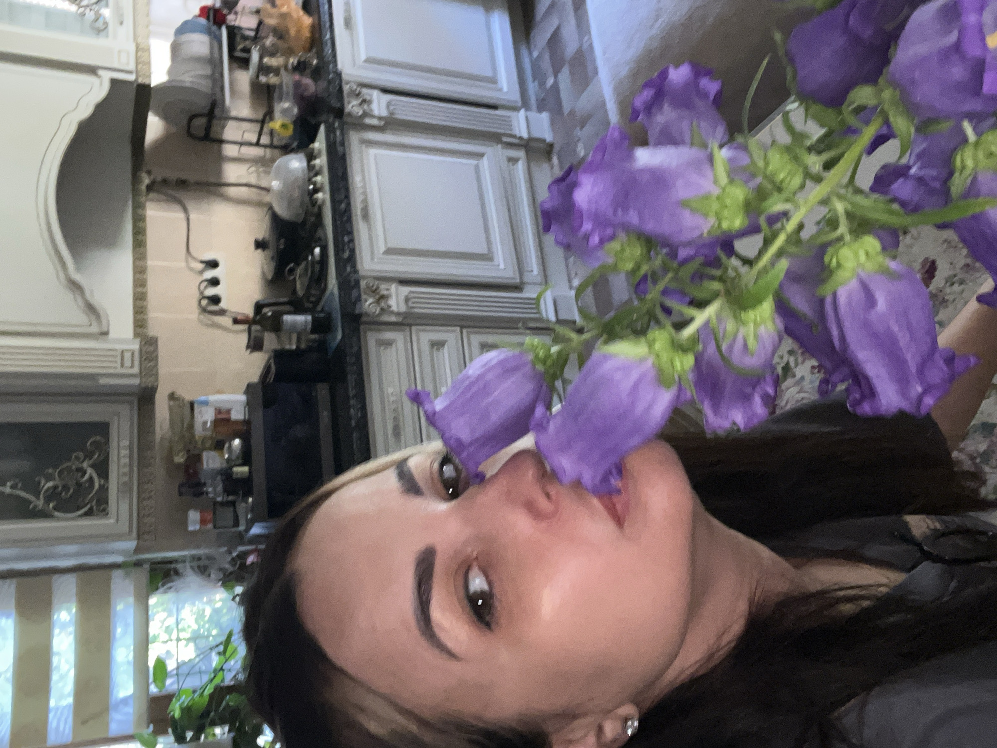 A woman is in a kitchen holding a bouquet of purple flowers. She is leaning in to smell the flowers, with her face partially obscured by them. The kitchen appears to be furnished with white cabinets, a dark countertop, and a microwave. The lighting is natural, coming from a window with blinds. The woman has dark hair and is wearing a dark shirt and earrings.