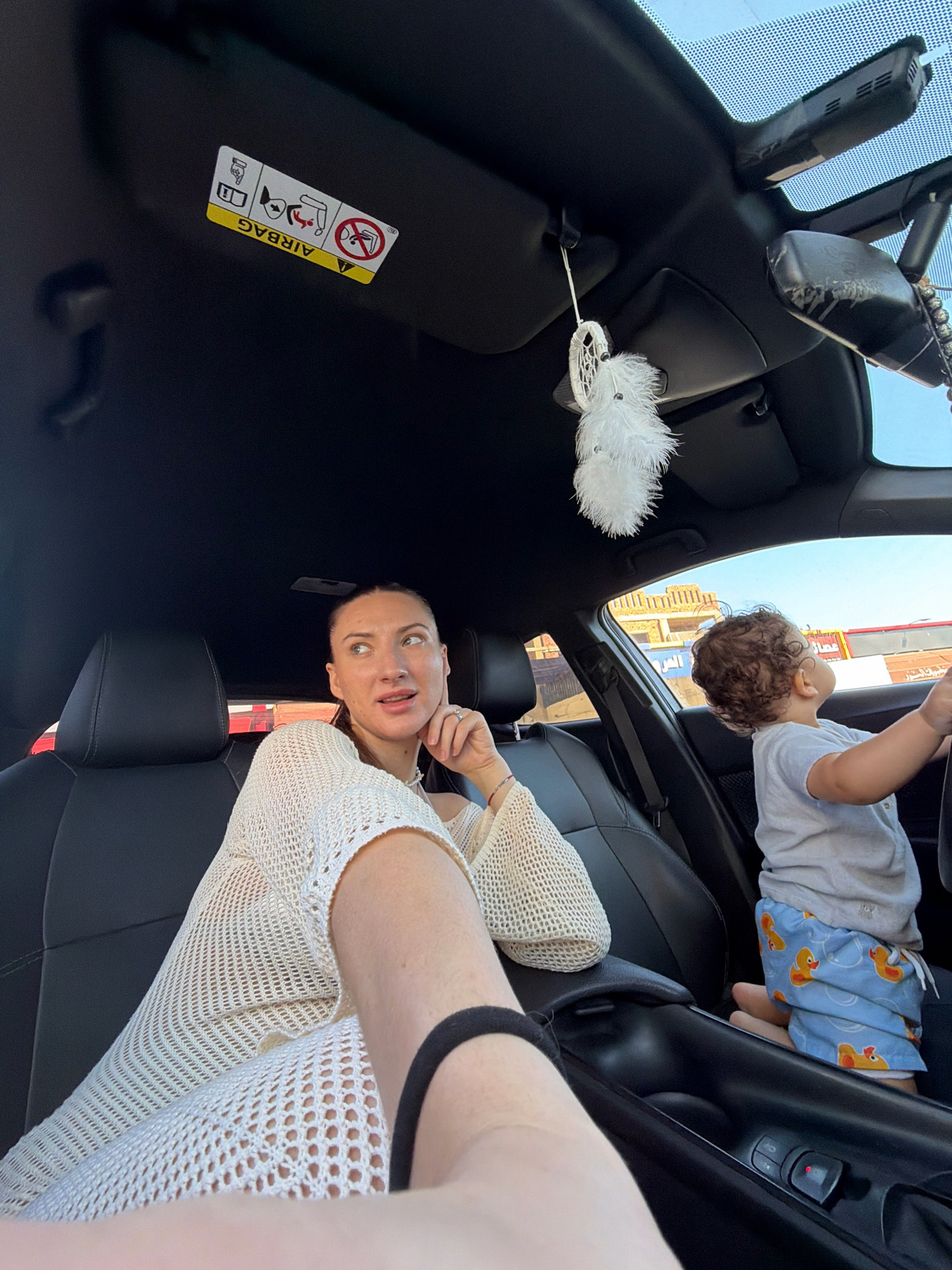 The image shows a woman and a baby in the front seats of a car. The woman is wearing a cream-colored, mesh-knit dress and is looking upwards and to the right, with her left hand resting on her chin. The baby, who is wearing a gray t-shirt and blue shorts with yellow duck patterns, is seated in the passenger seat, facing forward and appearing to hold the steering wheel. A white, fluffy ornament is hanging from the car's interior ceiling. On the driver's side sun visor, there is a yellow sticker with several symbols, including "AIRBAG" written in bold letters. The car's interior appears to be black leather. Outside the car, through the windshield and side windows, there are buildings and what appears to be a street scene, suggesting an urban or suburban setting. The bright sunlight and the architectural style of the buildings could indicate a location like Sharm El Sheikh, Egypt, where such arid and distinctive structures are common. The overall scene captures a candid moment of two individuals inside a vehicle, possibly during a journey.
