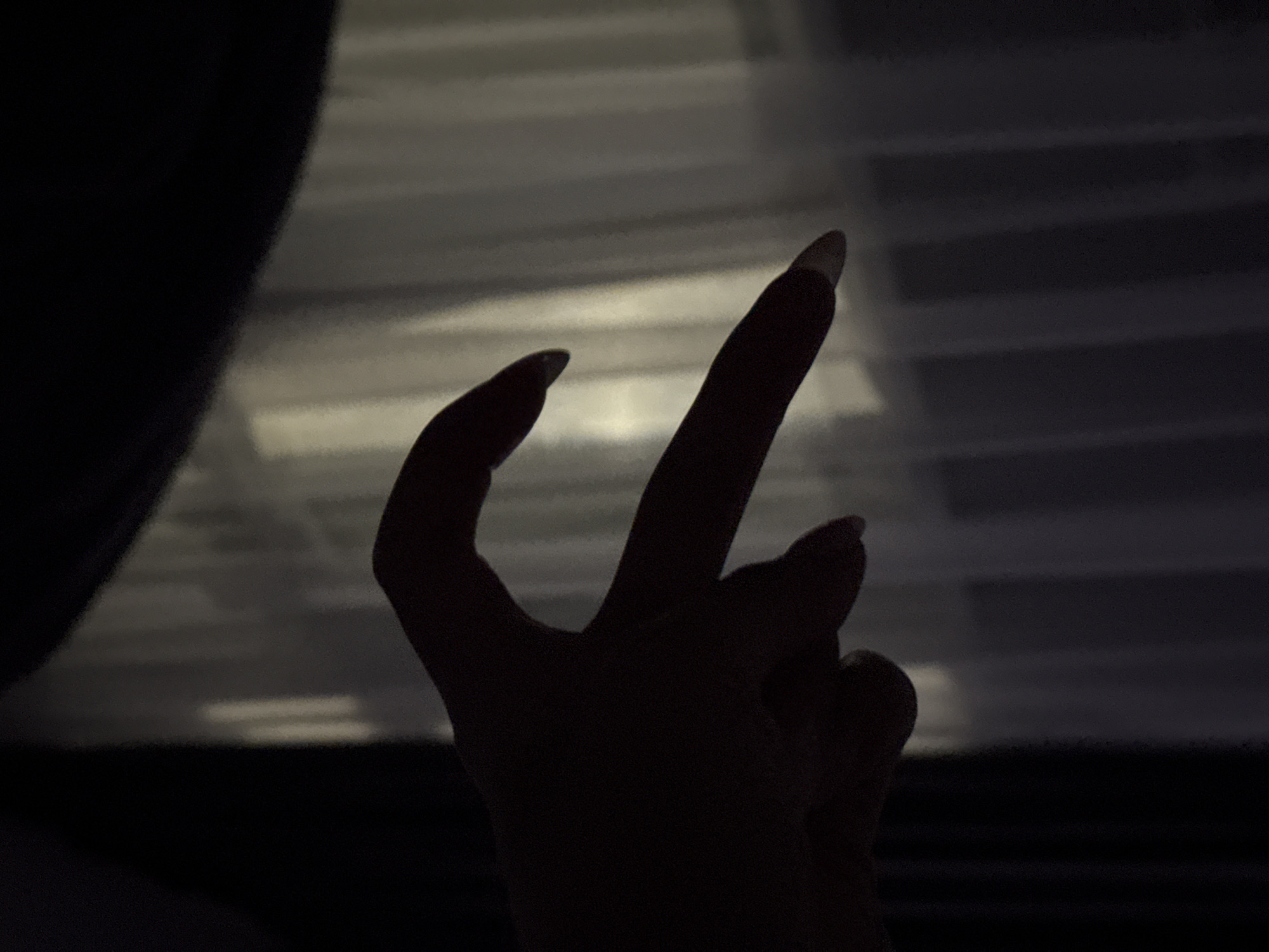 In the city of Doma 1096 Km, Russia, a close-up, silhouette photograph captures two hands forming a heart shape. The hands, positioned in the foreground, are silhouetted against a softly lit backdrop of vertical blinds. The left hand is curved inwards, with its thumb and index finger forming the top curve of the heart. The right hand's index finger extends and meets the curve of the left hand, completing the heart shape. The lighting creates a dramatic contrast, casting the hands in deep shadow while the blinds are illuminated by an unseen light source, suggesting an indoor setting with a window. The overall impression is one of intimacy and connection, conveyed through the gesture of the hands.